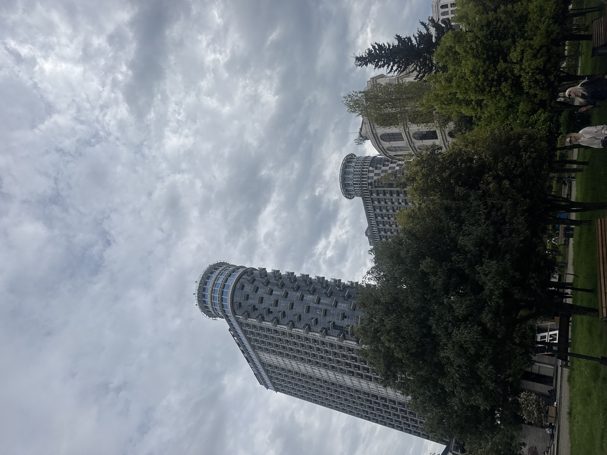 The image shows a tall building with a distinctive cylindrical top section, likely a skyscraper, viewed from below. The building is composed of many floors, with a repetitive pattern of windows. Trees partly obscure the lower section of the building. The sky is cloudy, and the lighting is subdued. In the foreground, there is green grass and benches. In the background, other buildings can be seen. The location is in a city but is unknown. People are also visible within the shot, but they're obscured in the background, so it's impossible to know what they are doing.