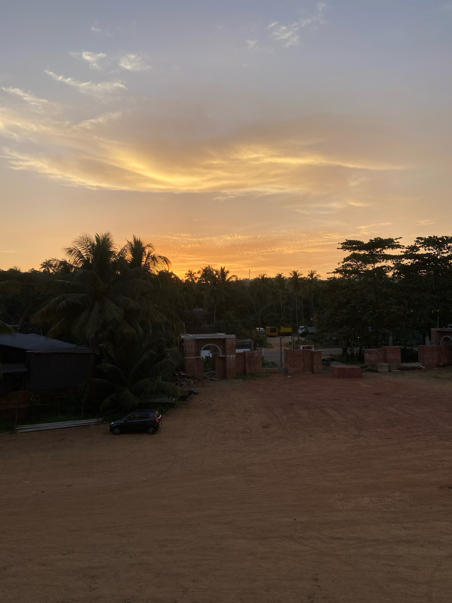 The image depicts a rural landscape at dusk, likely in the city of Malappuram, India. The sky is a canvas of warm hues, with streaks of orange and yellow clouds illuminated by the setting sun against a pale blue backdrop. In the foreground, a dirt road or cleared area dominates the scene, with scattered piles of red bricks and unfinished brick structures suggesting ongoing construction. To the left, a dark-colored, compact car is parked on the dirt. Adjacent to the car is a dark, shed-like structure. Beyond this, a dense grove of palm trees forms a lush green backdrop, interspersed with other trees. In the middle ground, a brick archway, also under construction, stands prominently. Further in the distance, more palm trees and other foliage are visible, with a few trucks and a white vehicle on a road. The overall impression is one of a developing rural area at the end of the day.