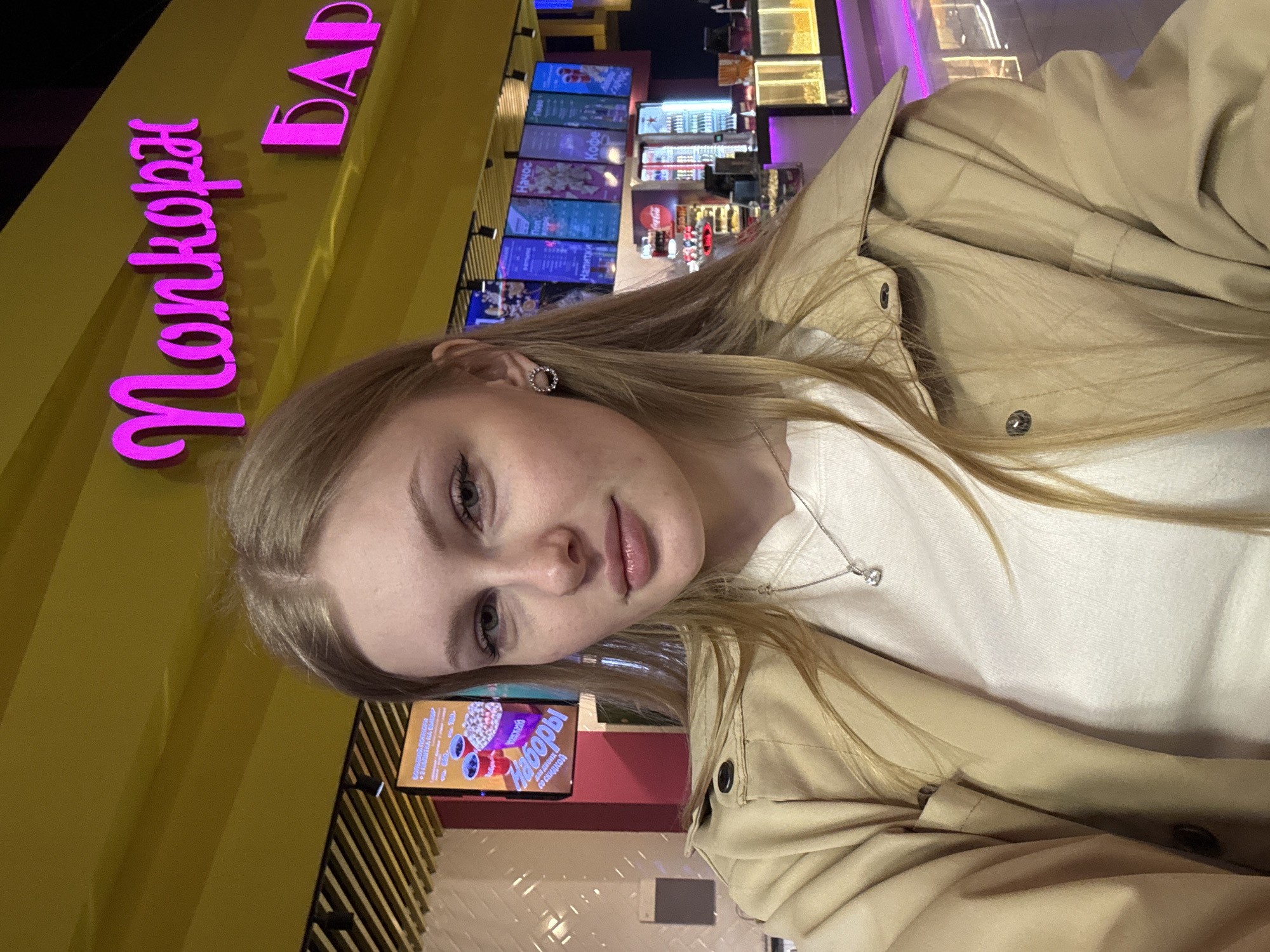 A young woman is standing at a concession stand in a movie theater in Perm, Russia, according to the text on the sign. She is wearing a beige jacket and a white t-shirt. She has blonde hair and is wearing earrings. The concession stand is selling popcorn and other snacks. There are digital screens showing the menu behind the counter.