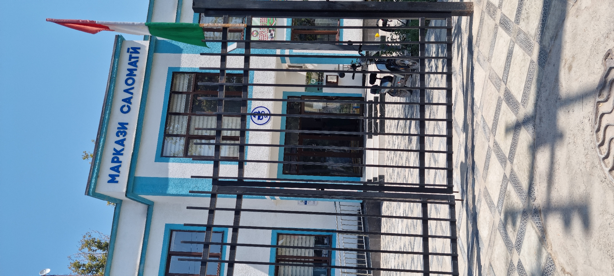 The image depicts the exterior of a building with a white facade and blue trim, identified by signage as "МАРКАЗИ САЛОМАТИ" which translates to "Health Center". A flag with horizontal stripes of red, white, and green is visible on a pole to the right of the building. The entrance features glass doors and steps, and a small electric scooter is parked in front of it. The building is enclosed by a black metal fence. The ground in front of the fence is paved with square tiles featuring a patterned design. The sky is clear and blue, and the scene appears to be bathed in sunlight, casting shadows from the fence and vegetation. The location is identified as the city of Ismoili Somoni, Tajikistan. There are no people visible in the image.