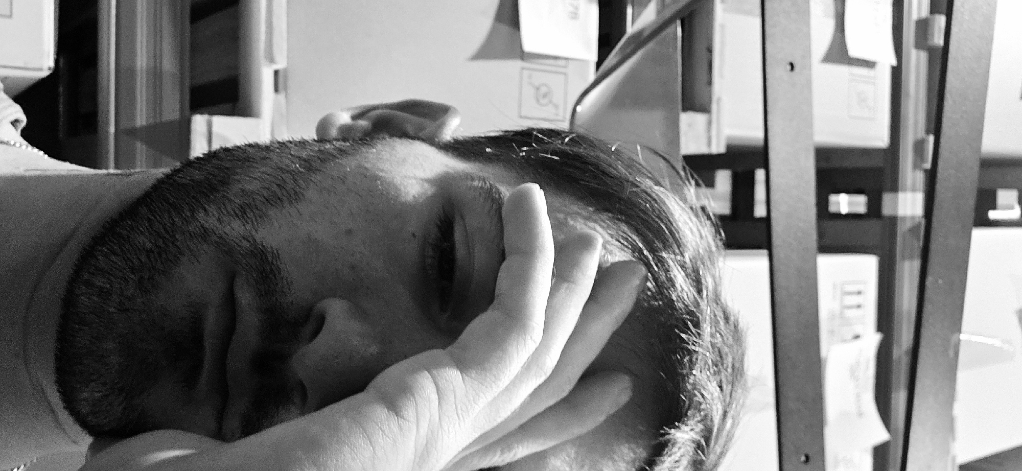 This black and white photograph presents a close-up, angled view of a person's head and hand, set in an indoor environment, possibly in Plovdiv, Bulgaria. The individual is positioned with their head tilted, their eyes seemingly closed or obscured, and one hand resting against their face, covering the lower part of their eye and nose. This posture suggests a state of sleep, deep rest, or possibly weariness and distress. The person has short, dark hair or stubble on the sides of their head, with longer hair visible on top. Their ear is also partially visible.
Strong, high-contrast lighting from the upper right illuminates parts of their face and hand while casting significant shadows, enhancing the monochrome aesthetic. The background, largely out of focus, reveals vertical dark structures resembling shelves or furniture supports. Various items, including white papers, labels, or small boxes, are discernible on these surfaces. Visible details include a symbol resembling a magnifying glass or an encircled 'a' on one white card, and faint numbers such as "176" and "111" on other background elements. The overall scene suggests a quiet, possibly solitary, moment of repose or exhaustion within what appears to be an office or storage environment. No specific time of day or weather is evident.