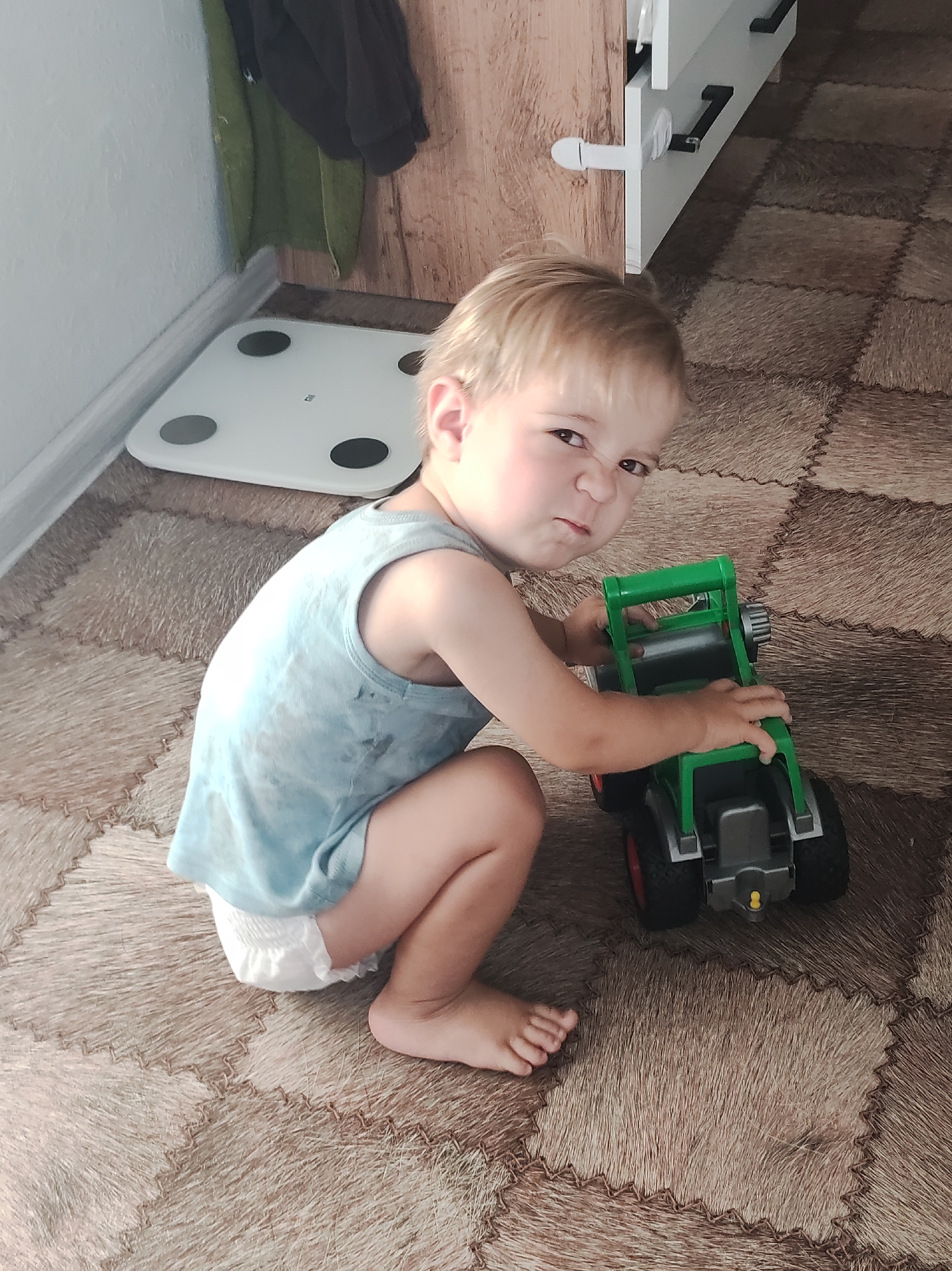 In an indoor setting, a young child with blond hair is crouching on a patterned floor, playing with a green toy tractor. The child, wearing a light blue tank top and a diaper, has a stern expression on their face. To the left of the child, a white digital scale is visible on the floor near a wall. In the background, a wooden surface and a piece of dark fabric are also present. The scene does not contain any specific indicators to place it in Chaikovsky, Russia.