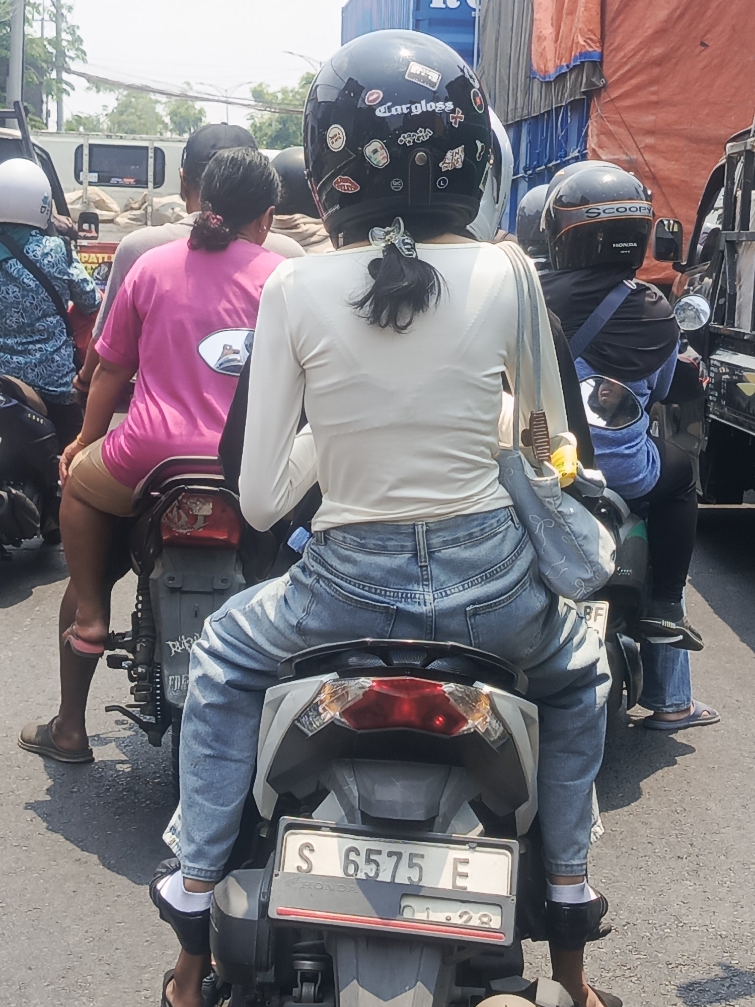 The image shows a busy street scene in Surabaya, Indonesia, with several motorbikes in traffic. In the foreground, a person wearing a white long-sleeved shirt and light blue jeans is riding a motorbike. The person has dark hair tied in a ponytail with a bow. The motorbike has a license plate that reads "S 6575 E" and "01.28". Another person wearing a pink t-shirt and beige shorts is on a motorbike behind the foreground rider. A third person wearing a black helmet with "Scoopy" written on it and a blue shirt is on a motorbike to the right. In the background, there are trucks and various buildings, suggesting an urban environment. The weather appears to be sunny.
