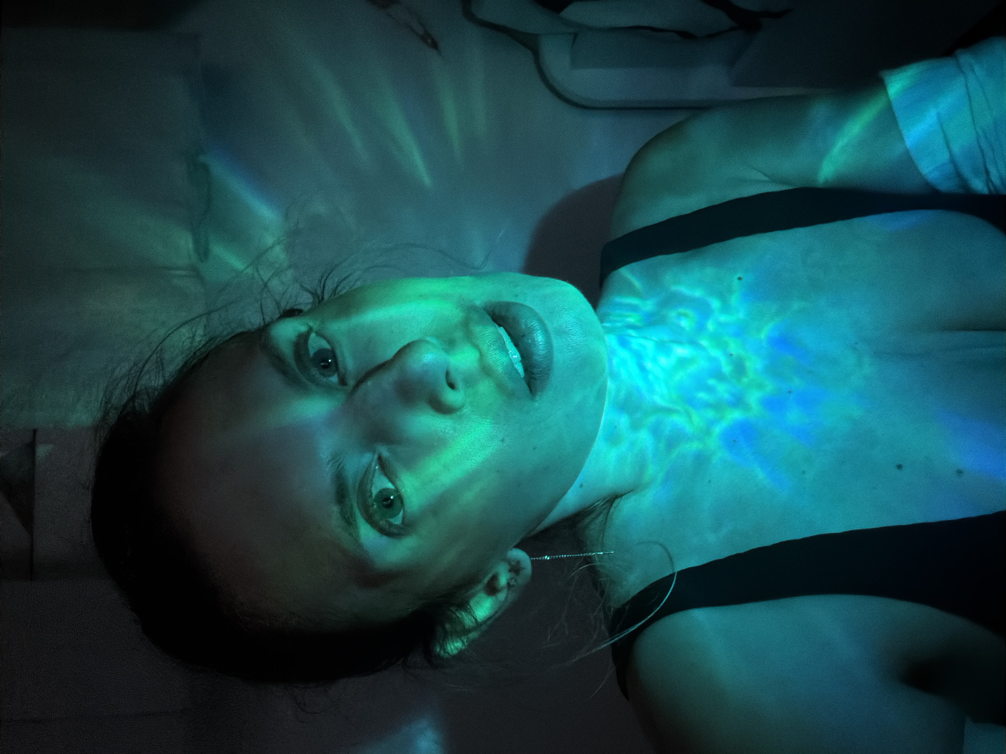A woman lies supine on a white surface, likely a bed. Her head is tilted back, and she is looking up toward the camera. She is wearing a black top. Her skin is illuminated by an unusual blue-green light pattern that resembles refracted light from water, particularly prominent on her chest, neck, and face. One can see a single silver earring hanging from her ear. The image is captured indoors. Location details such as the city of Budva, Montenegro cannot be inferred from the visible elements.