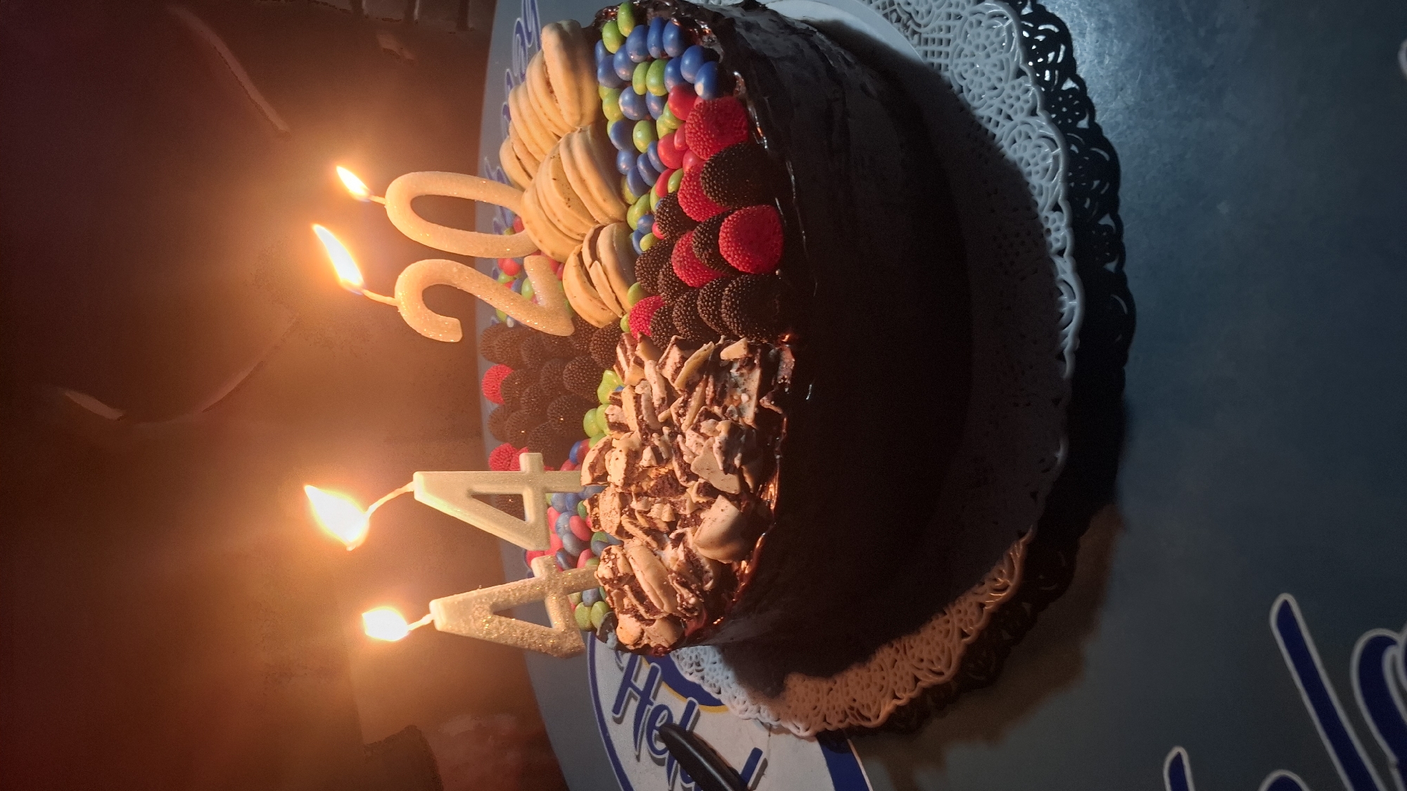 A birthday cake sits on a table, adorned with a variety of toppings, including white wafer rolls, M&M's, and assorted berries. Lit candles spelling out "44" and "20" are stuck into the cake. The cake rests on a white doily and a blue and white base with the partial words "Helader" visible. The image appears to be taken indoors, possibly in a home or restaurant in Buenos Aires, Argentina.