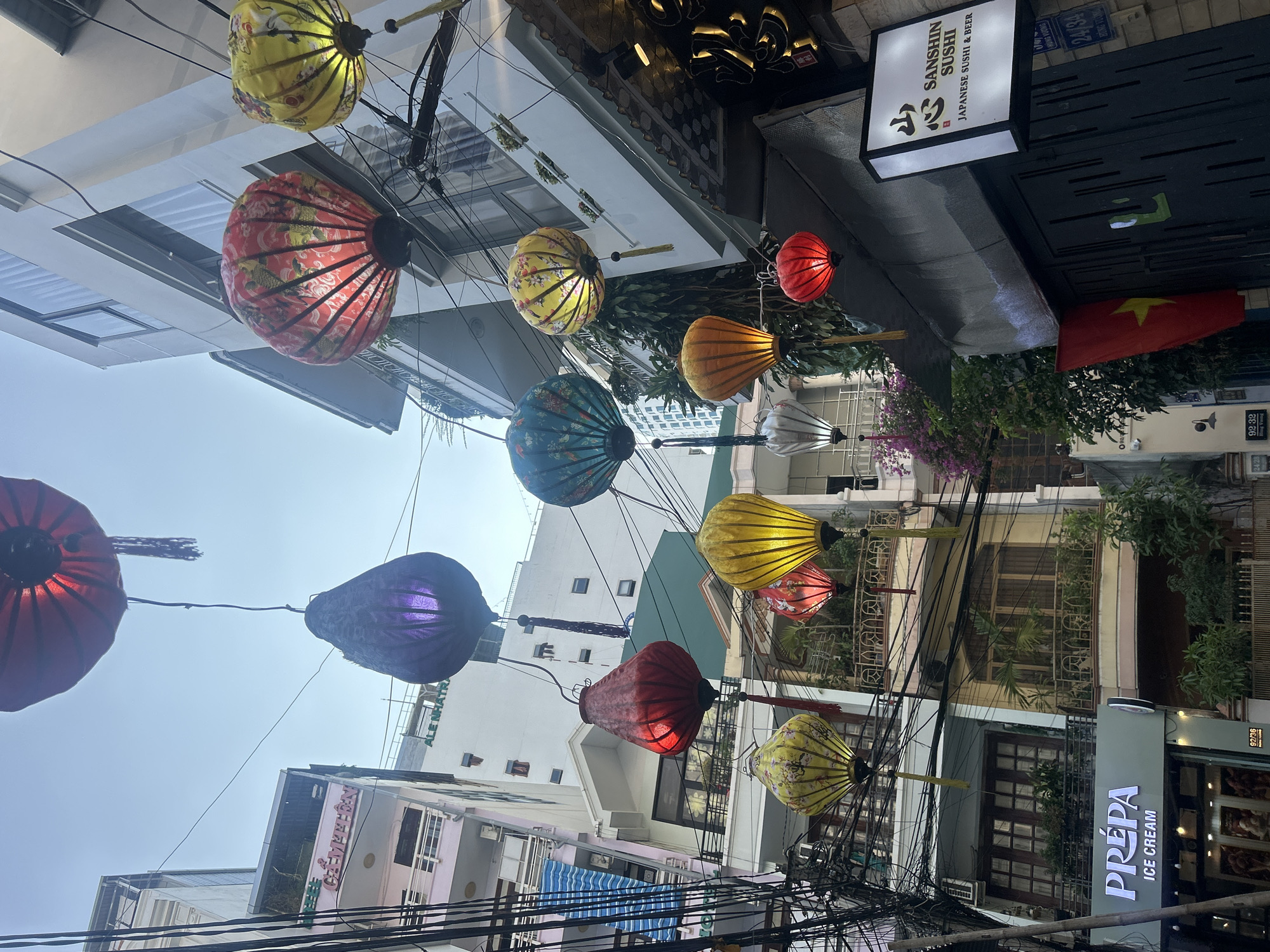 The image depicts a street scene in Nha Trang, Vietnam, characterized by colorful hanging lanterns strung across the street. These lanterns are of various sizes, shapes, and colors, including red, yellow, blue, orange, and purple. Overhead power lines crisscross the scene, adding to the visual complexity. Buildings line both sides of the street, with commercial establishments at street level. A Vietnamese flag is visible. Signs for "SANSHIN SUSHI JAPANESE SUSHI & BEER" and "PRÉPA ICE CREAM" are prominently displayed. Street numbers visible include 92/36, 24/28, 92/32, and 24/34. Vegetation, including flowering plants, adds touches of green and purple to the urban environment.