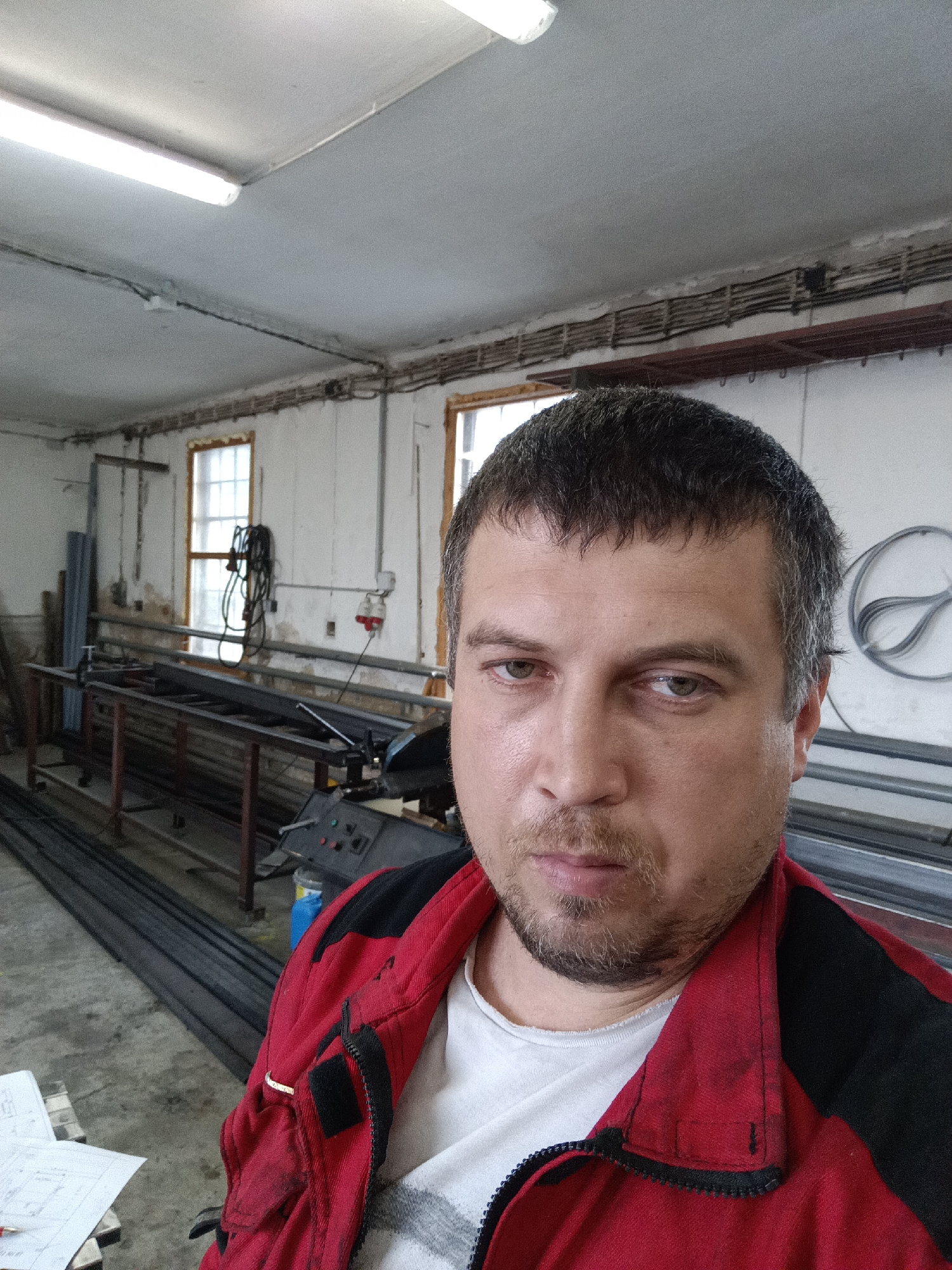 The image captures a man taking a selfie in an indoor workshop or industrial space in Dalovy, Czechia. The subject is a man, likely in his 30s or 40s, with short dark hair, light stubble, and light-colored eyes. He wears a red and black work jacket over a white t-shirt and looks directly at the camera with a neutral, somewhat serious expression.
The scene behind him is a functional workspace with white walls showing signs of wear and staining, a grey ceiling, and a concrete floor. A long fluorescent light fixture illuminates the ceiling, and exposed electrical wiring runs along the upper walls. Two multi-paned windows on the left wall allow natural daylight to enter, indicating it is daytime.
The workshop contains several metal workbenches or tables laden with stacked metal bars and profiles. A machine, possibly a metal cutting saw, is visible on one of the work surfaces. Other objects include a blue plastic container, an electrical outlet on the wall, and a coiled wire or metal object on the right side. Paperwork, possibly blueprints, is laid out on a table in the lower-left foreground, though no specific text is legible. The environment suggests an active, utilitarian work setting.