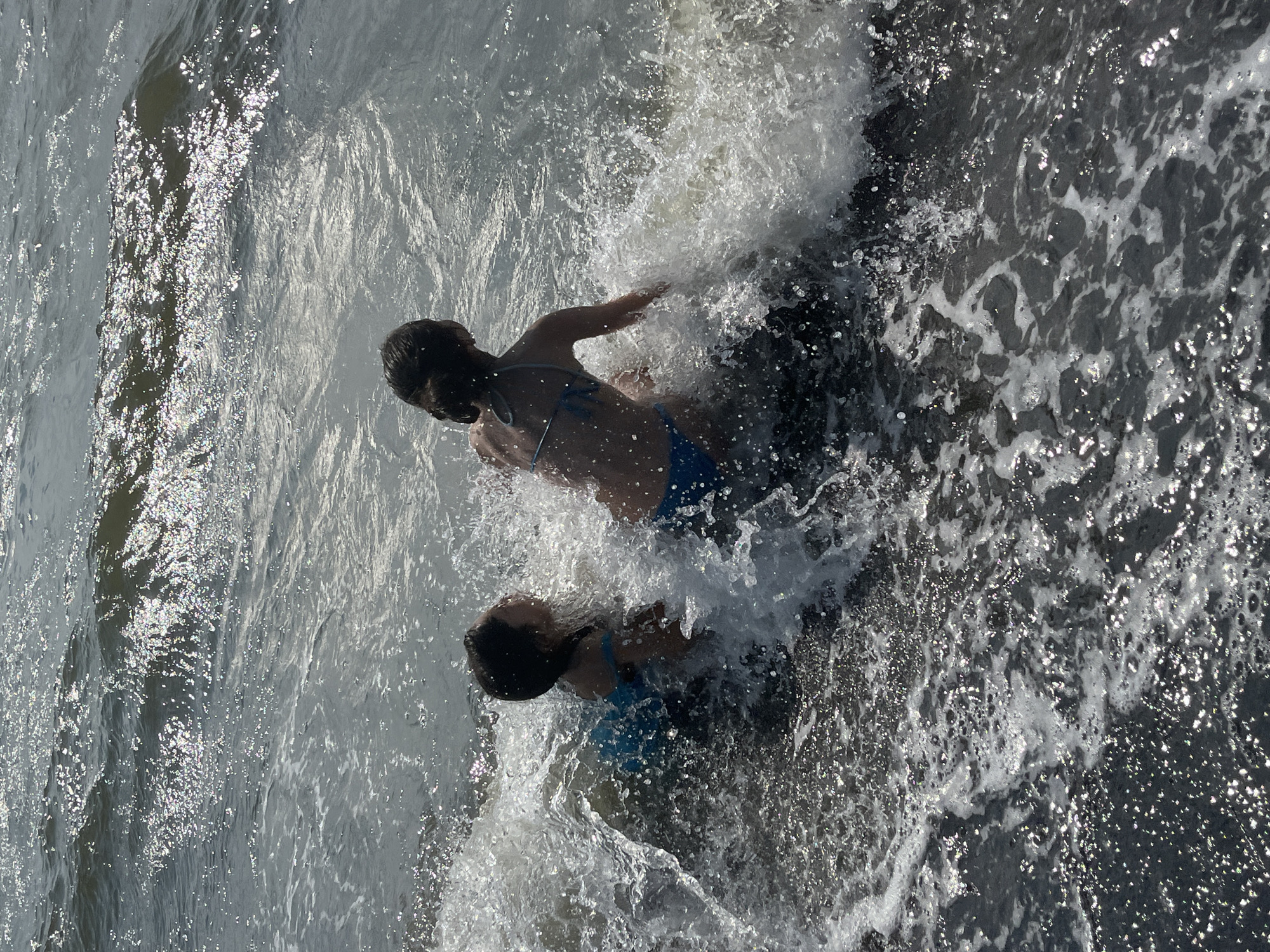 This media file shows two individuals playing in the ocean. They are both in the water, with waves splashing around them. The person in the foreground is wearing a blue bikini and is positioned lower in the frame, appearing to be submerged slightly. Their head is visible, with dark hair. The second person is above and to the left of the first, also with dark hair, and is more visible, appearing to be on their stomach with their arms outstretched, possibly swimming or playing in the waves. The water is choppy and bright, with sunlight reflecting off the surface, suggesting it is a sunny day. The scene is a beach or coastline, likely Ureki, Georgia, given the context. The activity appears to be casual and recreational, with a sense of enjoyment and playfulness suggested by the splashing water. There is no visible text.