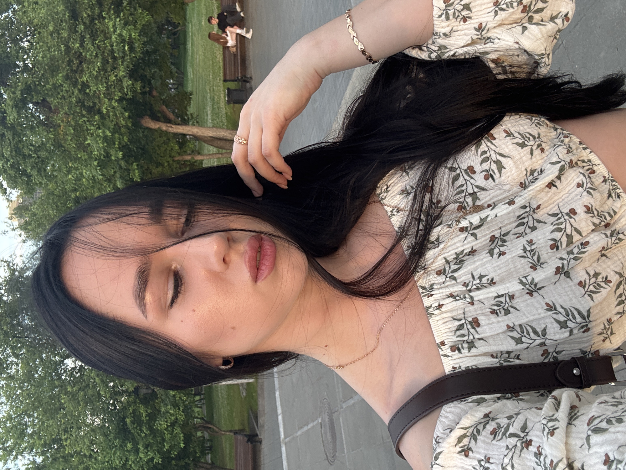 A young woman with long, dark hair is captured in a close-up, outdoor shot, likely taken in a park in Novosibirsk, Russia. Her eyes are closed, and her head is tilted down, with strands of hair falling across her face. She is wearing a cream-colored off-the-shoulder top with a floral pattern. A thin gold necklace rests on her collarbone, and she has a delicate gold ring on her right ring finger and a matching bracelet on her right wrist. Her skin appears to be glowing in the soft, natural light, suggesting it might be either sunrise or sunset. In the background, trees with lush green foliage frame the scene, and park benches are visible, with two other individuals seated on one of them in the distance, their figures blurred. The ground appears to be paved, possibly a walkway or plaza.
