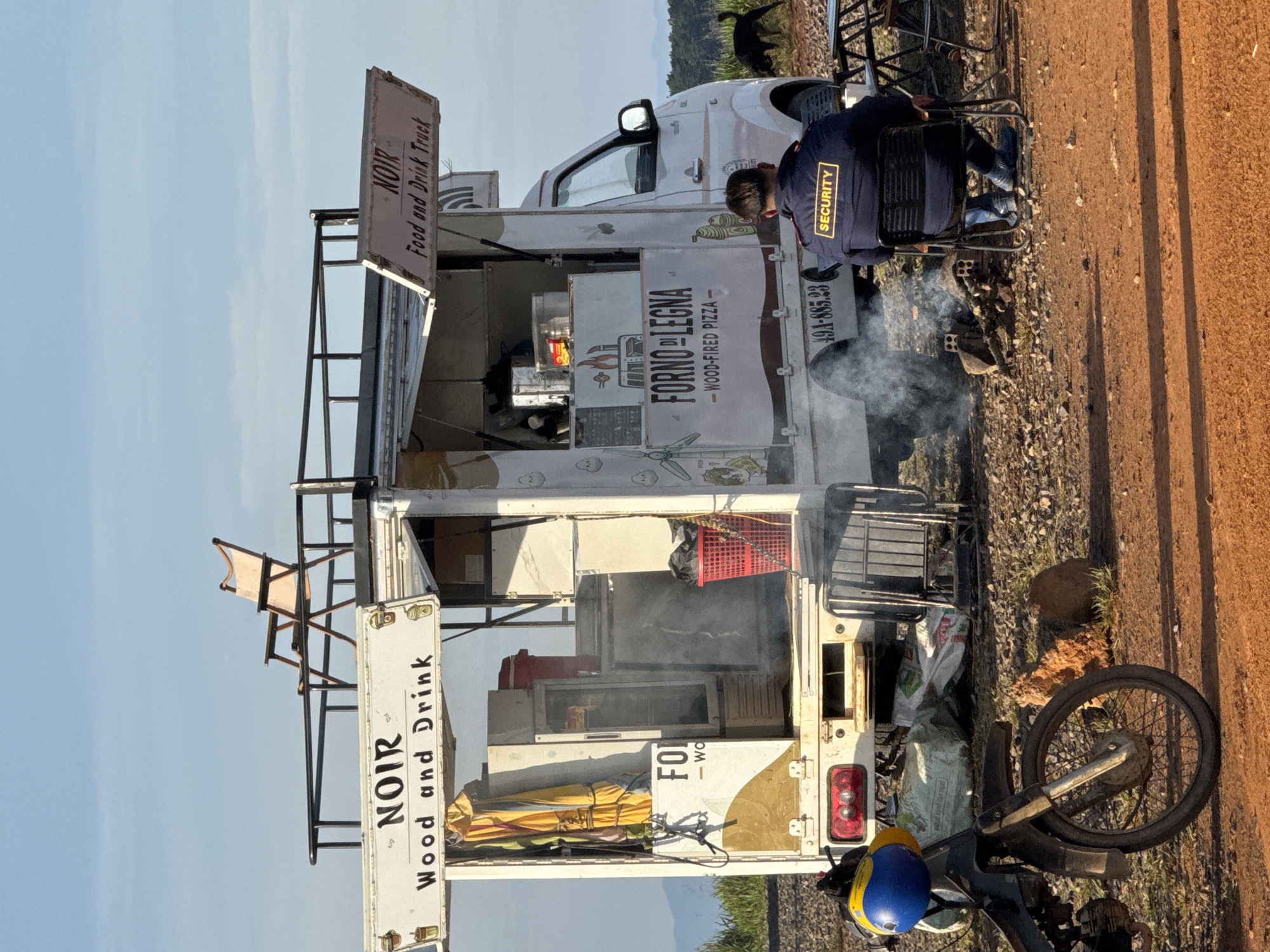 The image depicts a food truck named "NOIR Wood and Drink" and "NOIR Food and Drink Truck" parked on a dirt road in Trường Thọ, Vietnam. A security guard, wearing a dark blue jacket with a yellow "SECURITY" patch on the back, sits in a black folding chair facing away from the camera. In front of the security guard, a small fire burns, producing smoke. Adjacent to the food truck is a motorcycle with a blue and yellow helmet. A black dog is visible in the background, near a table and chairs set up outdoors. The food truck itself is white with colorful decals. One decal advertises "FORNO DI LEGNA - WOOD-FIRED PIZZA." Above the truck, a light pink folding chair is placed on a raised platform. The overall setting appears to be an open area with sparse vegetation and distant hills under a clear blue sky.