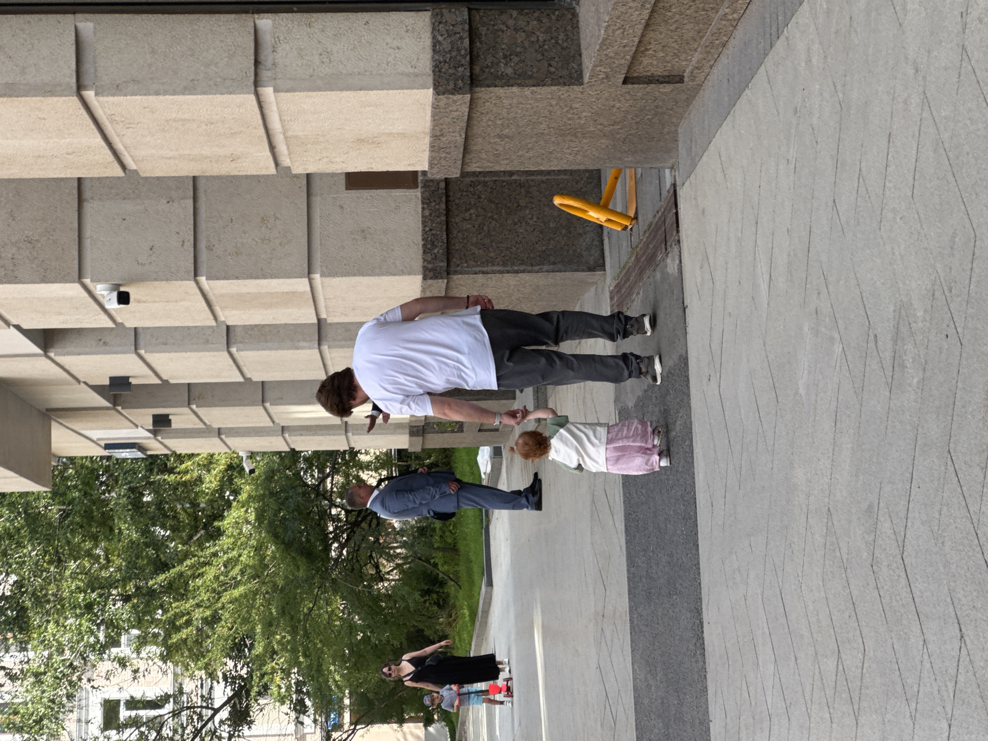The image depicts a group of people in an outdoor urban setting, likely Moscow, Russia, on a bright, sunny day. The scene appears to be a sidewalk or plaza adjacent to a building with a textured stone facade.
The main subjects are several individuals. In the foreground, a woman in a black dress is visible, with two children near her, one of whom is on a scooter. Further back, a man in a blue suit stands. In the mid-ground, a man in a white t-shirt and grey pants is holding the hand of a young child, who is wearing a pink skirt and a white top. The interaction between the man and child suggests a moment of connection or guidance.
The activity appears to be a leisurely stroll or outing. The presence of children on a scooter and the generally relaxed postures of the adults imply a casual atmosphere.
Notable details include the architecture of the building, which features large, square stone panels. A yellow object, possibly a bike rack or a piece of street furniture, is visible in the background. The lighting suggests it is daytime, with clear skies contributing to the bright and possibly warm weather. The overall impression is one of everyday urban life. No visible text is present in the image.