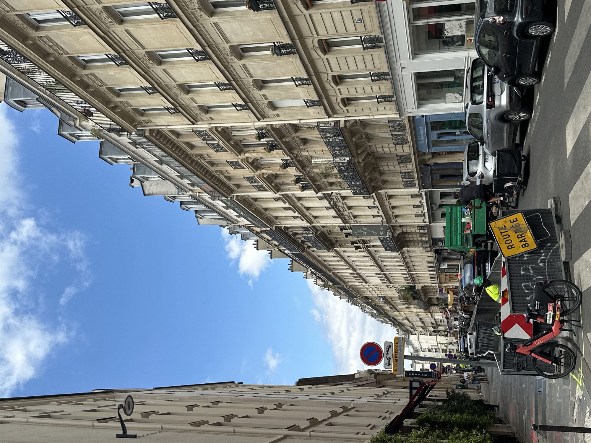 This is a tilted, eye-level, medium shot of a Parisian street scene with buildings lining the right side of the frame and a bright blue sky with scattered white clouds filling the left. The buildings are made of light-colored stone and feature multiple stories with ornate balconies and regularly spaced windows.
On the street, there are several cars parked along the sidewalk, including a black SUV and a silver sedan. In the foreground, a red and black shared bicycle is parked. A person wearing a bright yellow safety vest and green helmet is visible near some construction equipment and a green truck. There are also various signs visible: a yellow sign with black text reads "ROUTE BARRÉE," indicating a closed road. A red circular sign with a blue bar and a red diagonal line through it signifies "no parking." Another yellow sign with black text says "À COMPTER DU."
The overall impression is of a sunny day in a city, likely Paris, given the architectural style and the presence of shared bicycles and signage typical of European cities. The activity appears to be ordinary street life, possibly with some temporary disruption due to road work or an event indicated by the "ROUTE BARRÉE" sign. The sunlight suggests it's daytime. The image is captured from a slightly low angle, looking up at the buildings and the sky.