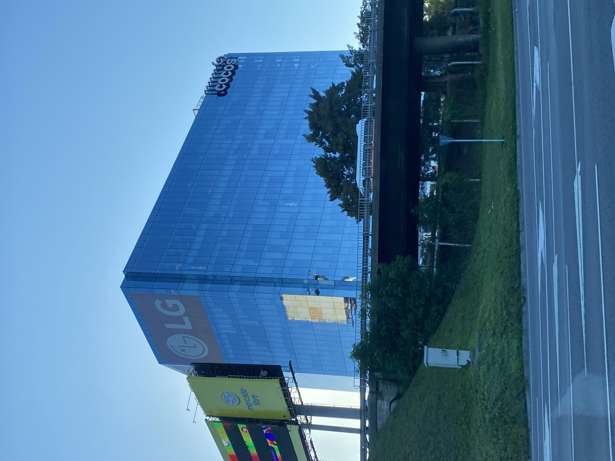 The image shows a tall, modern glass-fronted building in Vicente López, Argentina. The building displays the "Telecom" logo at the top, an LG logo lower down, and a billboard advertising "Mercado Libre" at the bottom. A roadway and grassy area are visible in the foreground. An elevated structure, possibly a bridge or overpass, passes close to the building. The sky is clear and blue.