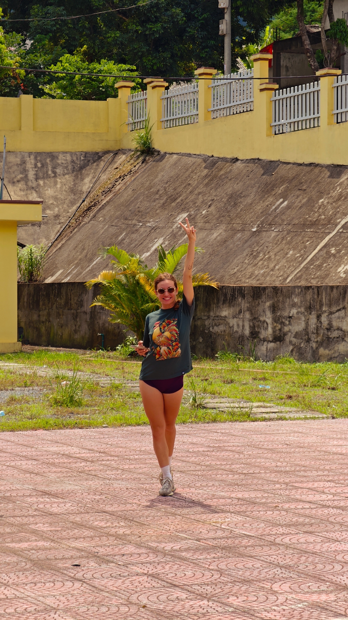 A woman is captured mid-stride, smiling and raising her left arm to make a peace sign (V-sign) with her fingers. She holds a smartphone in her right hand. She has light brown hair, wears sunglasses, a dark green t-shirt featuring a colorful graphic of two roosters, dark purple athletic shorts, white socks, and light-colored sneakers. She appears happy and relaxed.
The woman is walking on a reddish-pink tiled ground with an intricate circular pattern. Behind her, a patch of overgrown grass and scattered concrete slabs separate the foreground from the background elements.
The background features a prominent yellow concrete wall topped with a white picket fence, extending horizontally across the image. Behind this wall, a large, upward-sloping concrete embankment is visible, partially covered with sparse vegetation and dirt. Lush green trees and tropical plants, including palm trees, fill the upper and mid-background, suggesting a verdant environment. A small part of a yellow building is visible on the far left.
The scene is brightly lit by natural daylight, indicating a sunny day. The overall impression is one of a cheerful moment in an outdoor, somewhat rural or undeveloped setting in Mường Kim, Vietnam. There is no visible text.