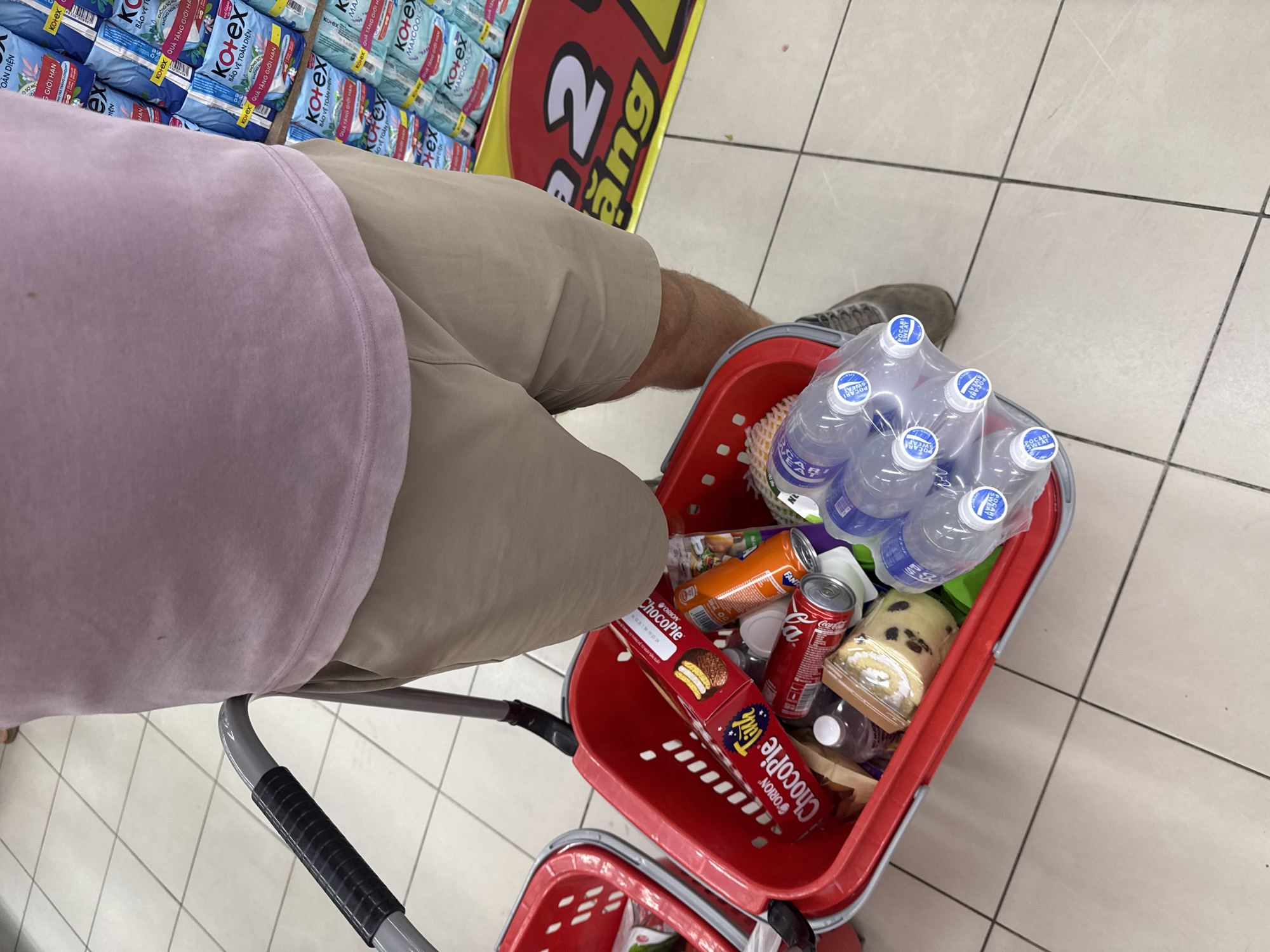 A person wearing a pink t-shirt and beige shorts is standing in a supermarket aisle, pushing a red shopping basket. The basket contains several items, including a box of ChocoPie cookies, a can of Coca-Cola, an orange soda can, a rolled cake with raisins, and several bottles of Pocari Sweat. In the background, shelves are stocked with Kotex feminine hygiene products. The floor is tiled with a white and beige checkerboard pattern. The scene appears to be in a store in Đông Hoa Lư Ward, Vietnam, as indicated by the Vietnamese text on the product packaging and store signage.