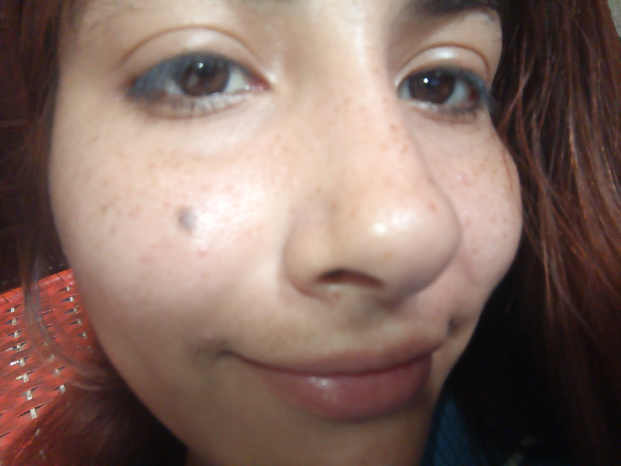 A close-up photograph shows a woman's face. She has fair skin with numerous freckles, and a mole is visible on her left cheek. Her eyes are brown and she appears to be wearing blue eyeliner. The woman has a slight smile. Reddish-brown hair is visible on the right side of the frame. A red woven object is located on the lower left corner. There is no other information, and the location in Juan Griego, Venezuela is assumed information not present in the image.