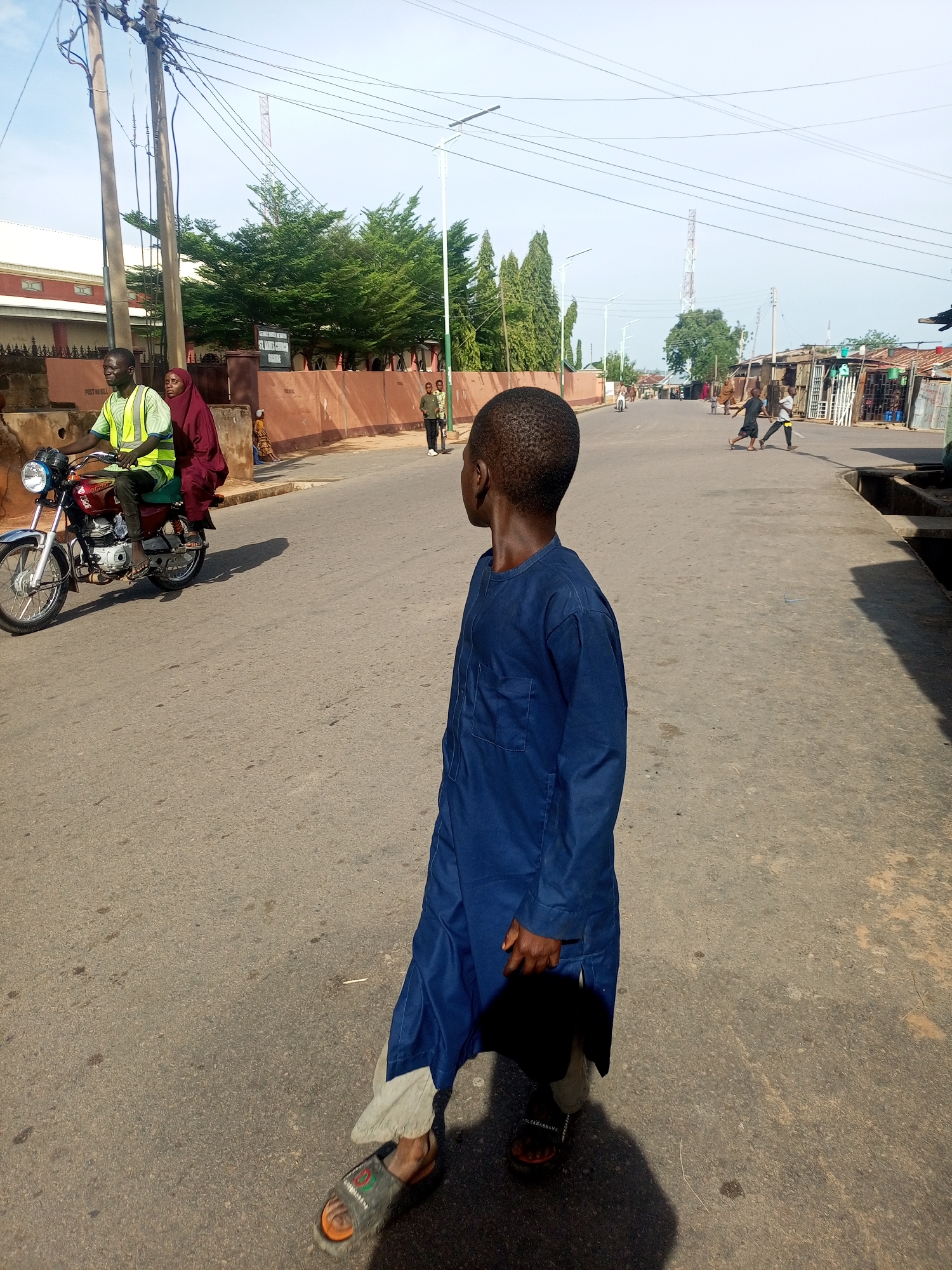 The image displays an outdoor street scene under clear daylight, located in the city of Unknown, Unknown. In the lower-right foreground, a young Black boy, estimated to be between 6-10 years old, stands with his back partially to the camera, looking left towards the middle distance. He is dressed in a long-sleeved, loose-fitting blue tunic over lighter trousers and wears open-toed sandals. He holds a dark object in his right hand, casting a distinct shadow on the asphalt road.
To the left of the foreground boy, a Black man rides a red and silver motorcycle towards the camera's left, wearing a bright yellow-green reflective vest over dark clothing and sandals. A Black woman, wearing a dark red hijab and matching attire, is seated as a passenger behind him. Behind the motorcycle, several utility poles with numerous overhead wires are visible. A reddish-brown wall with "POST NO BILL" visible on its surface runs parallel to the street, separating it from a multi-story building with a light roof and reddish walls, partially obscured by green trees.
Further down the asphalt road, in the midground, two additional young Black boys are observed walking or playing in the street. On the right side of the road, a concrete drainage ditch runs parallel to the pavement, beyond which are structures resembling informal market stalls or shops with corrugated metal roofs.
The background features a broad, light blue sky traversed by overhead power lines. A tall, slender lattice telecommunication tower is prominently visible in the distant center. Further greenery and modern streetlights are dispersed throughout the background, indicative of a semi-urban environment.