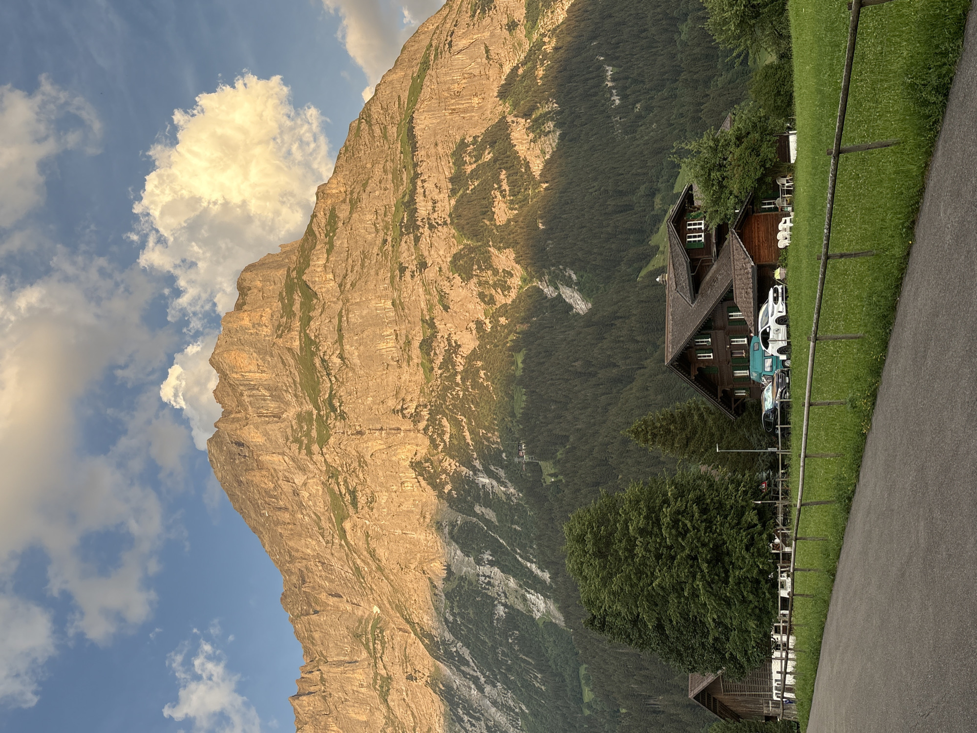 The image depicts a striking mountain landscape with a traditional wooden house and a grassy area in the foreground. The dominant subject is a massive, rugged mountain face, bathed in the warm glow of the setting sun, casting long shadows across its rocky surface. Patches of green vegetation cling to the mountain's lower slopes.
In the middle ground, a charming, dark wooden house with a steep, gabled roof and white-framed windows is nestled against the trees. The house appears to be a dwelling or perhaps a guesthouse. In front of the house, several white plastic chairs are arranged, suggesting an outdoor seating area. White vehicles, including what looks like a camper van, are parked nearby.
A neatly mown, bright green lawn stretches from the house towards a wooden fence that borders a paved path or road in the immediate foreground. The sky above is a vibrant blue, dotted with fluffy white clouds, one of which is illuminated by the sun. The lighting indicates it is late afternoon or early evening.
The scene evokes a sense of peacefulness and natural beauty, typical of alpine settings. The location is implied to be Grindelwald, Switzerland, given the context provided. There is no visible text in the image.