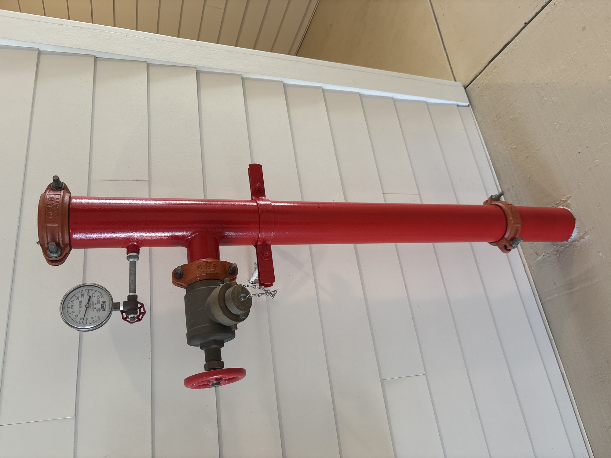 A red fire hydrant is mounted on a white vinyl-sided wall in Palmetto, United States. A pressure gauge is attached to the hydrant near the top. A valve with a red handle is located on the side of the hydrant. The hydrant is anchored to the wall by a bracket. The hydrant is positioned on a concrete surface. The scene is well-lit and the details are clear.