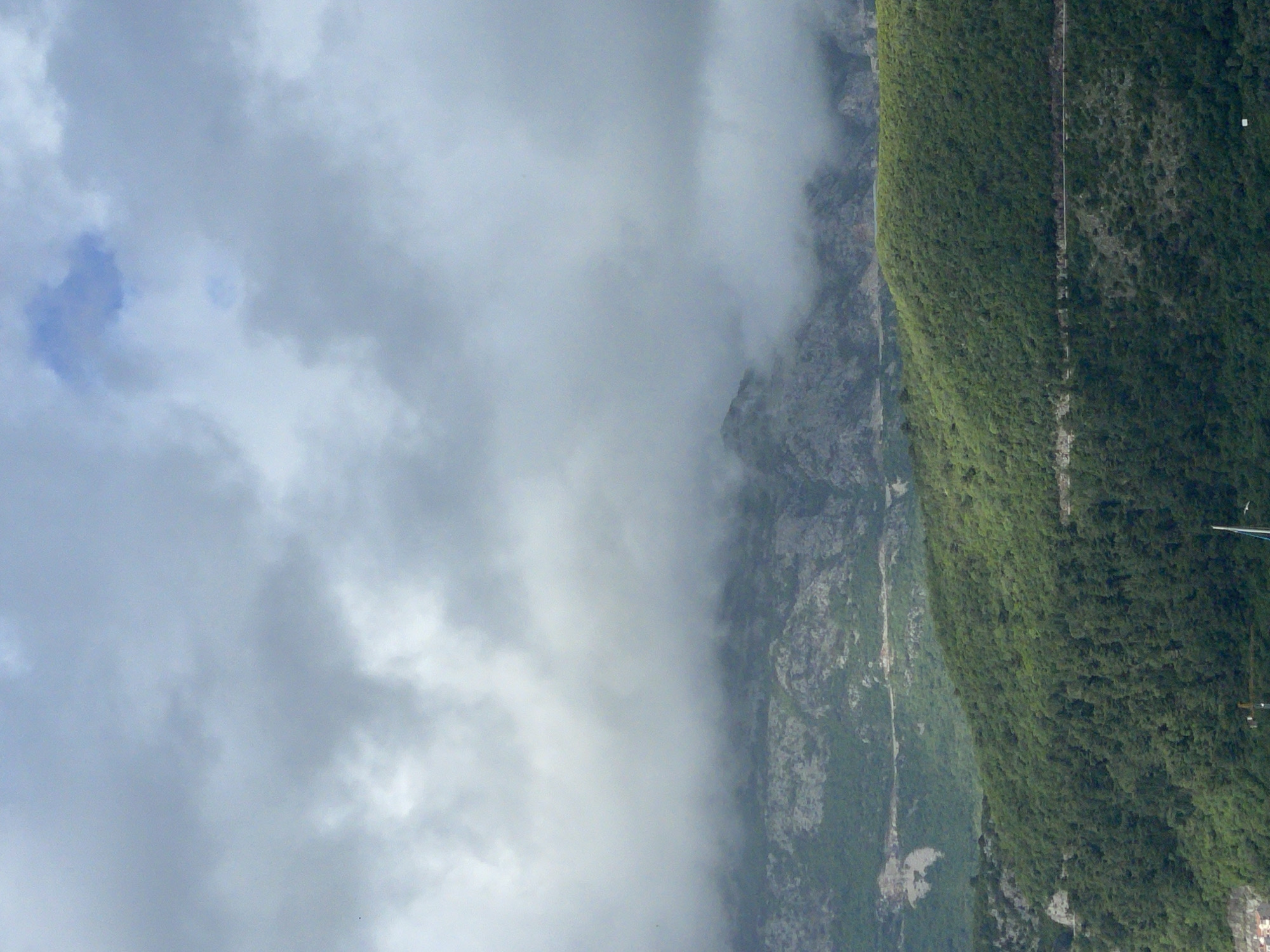 The image shows a mountain partially obscured by clouds. The right side of the mountain is covered in dense green vegetation. A narrow, light-colored path or trail can be seen running vertically along the mountain. The left side of the mountain is composed of rocky terrain that fades into the clouds, making it difficult to discern the exact features. The sky is filled with gray and white clouds, with a small patch of blue sky visible in the upper left corner. The location appears to be a mountainous area.