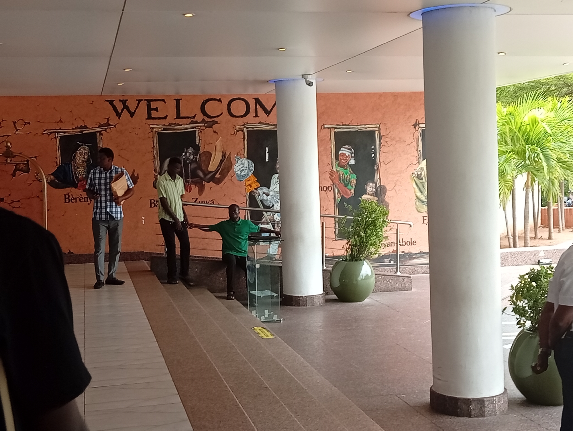 **Environment:**
The scene takes place in a covered outdoor area, possibly the entrance or lobby of a building. The floor is composed of light-colored tiles and dark tile accents, and there are steps leading up to a raised area. Tall white cylindrical columns support the overhead structure. The wall is painted in shades of orange and brown, with large mural-style depictions of people. Decorative potted plants are present.
**People:**
* Several people are present in the scene.
* One man in a plaid shirt and light-colored trousers is walking.
* Another man in a light green shirt and dark trousers is standing next to stairs.
* A man in a green shirt is standing with his arms outstretched.
* A person is partially visible on the left side of the frame wearing a black shirt.
* Another person is partially visible on the right side of the frame wearing a white shirt.
**Objects:**
* A glass barrier is present near the steps.
* A yellow sign is affixed to the step.
* The wall behind the people has the word "WELCOME" painted on it.
* Murals showing various individuals and scenes are painted on the wall.
**Interactions:**
The people appear to be in transit or waiting within the building's entry space. There is no obvious interaction between them.
**Location:**
The wall contains murals of the letters "Berény", "Zinvà", "E" and "an_bole" on the wall which suggest the location might be in Itirin, Nigeria.