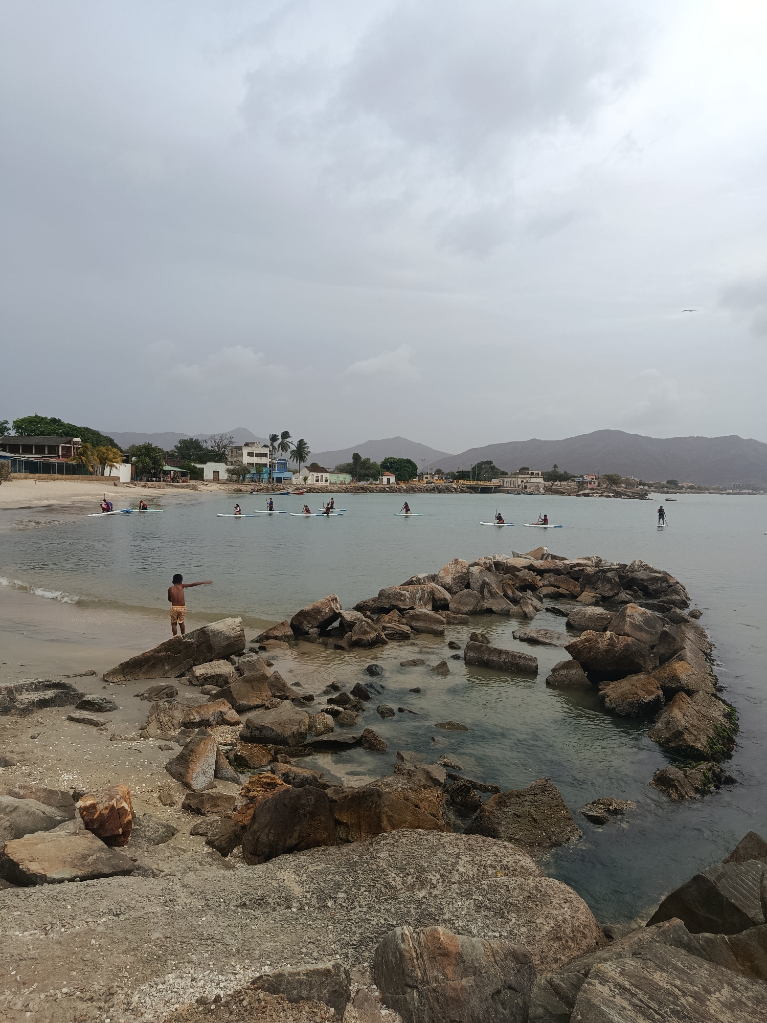 The image depicts a beach scene in Juan Griego, Venezuela, under an overcast sky. A stone breakwater extends into the calm water. A boy stands on the rocks near the beach, gesturing towards a group of people paddleboarding in the bay. In the background, there are buildings and low mountains visible along the coastline.