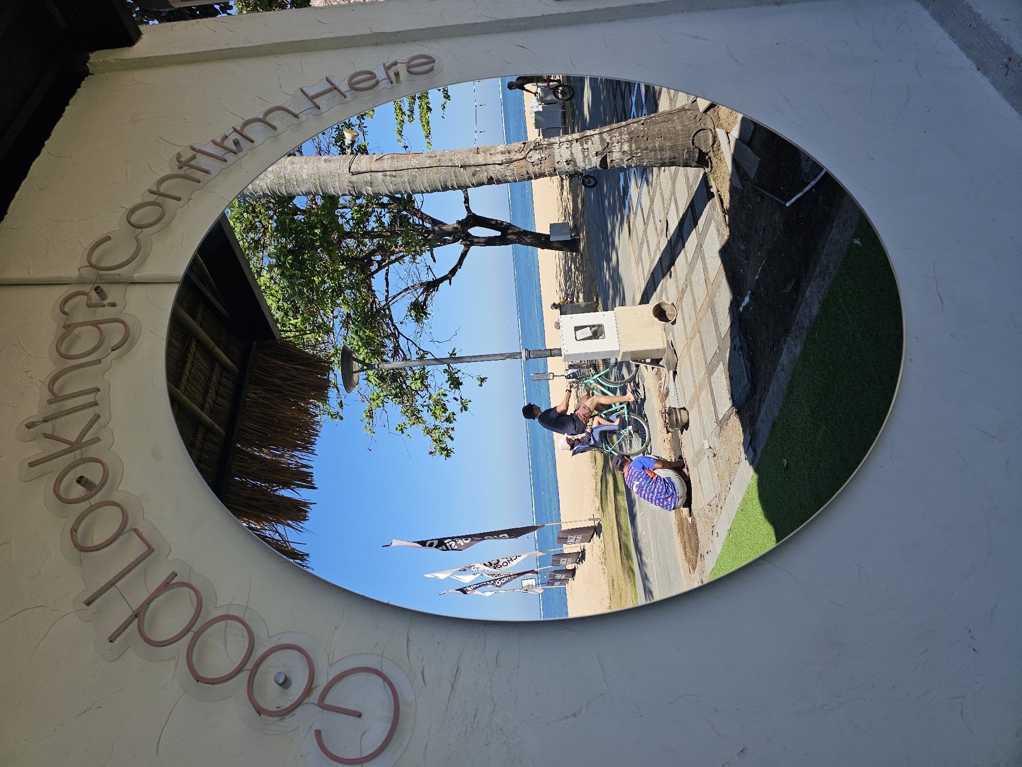 The image shows a framed view of a beach scene in Denpasar, Indonesia. The frame is a white archway with pink neon lettering that reads "Good Looking? Confirm Here".
In the reflection within the archway, a beach is visible with the ocean in the background. The sand is light brown and there are several black and white flags on poles planted in the sand. A man is riding a light blue bicycle with a child seated behind him. In the foreground of the reflection, a person is kneeling on the ground, appearing to be working on paving stones or the ground. A bucket is next to them. A palm tree is visible to the right, and a leafy green tree is to the left, casting shadows on the paved area. The sky is clear and blue.