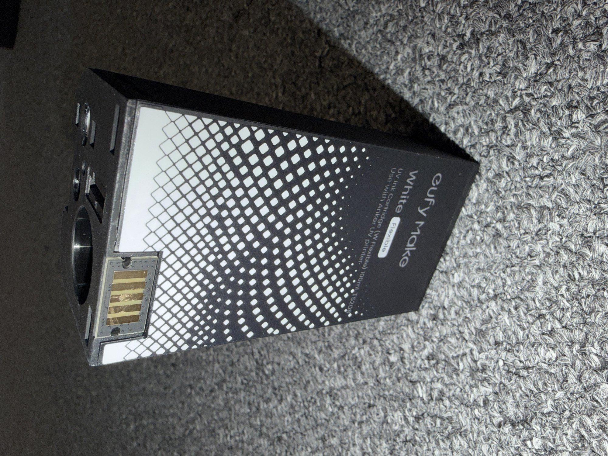This is a close-up, high-angle shot of a black product box with a white, geometric pattern on its side. The box is lying on a textured gray carpet. The text on the box reads "eufy Make White Flexible UV Ink Cartridge (W Flexible) 100ml T5208 Use with Anker UV printer." On the left side of the box, there is a metal connector with visible gold-colored pins. The lighting suggests it is indoors, possibly in a home environment, and no specific time of day or weather conditions are discernible. The image focuses solely on the product and its packaging, with no people or active event depicted. The overall impression is of a product shot intended to showcase the item and its labeling.