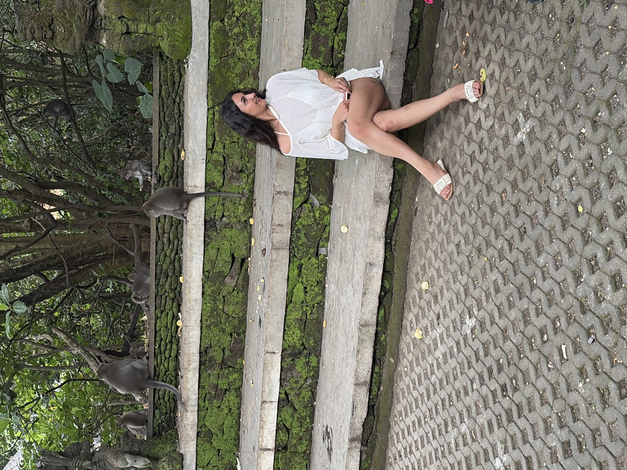 A woman is sitting on a stone step in what appears to be a park or temple complex in Ubud, Indonesia. She is wearing a white cover-up over a white bikini. The steps are covered in moss and surrounded by trees. Several monkeys are visible in the background, some sitting on a stone wall and others in trees. The ground in front of the steps is paved with interlocking concrete blocks. The woman is looking to her right and smiling.