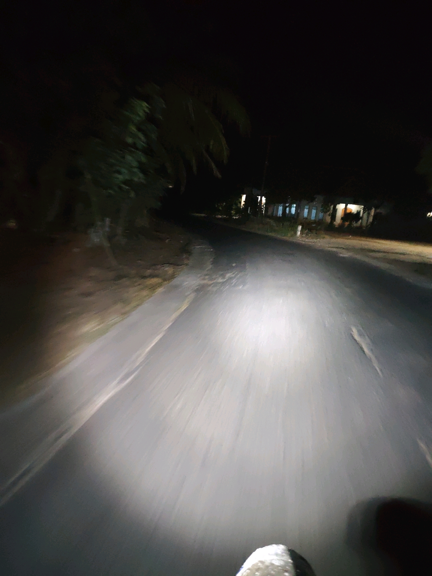 The image shows a first-person perspective of riding a motorcycle at night on a paved road in Penantian, Indonesia. The road is dark, with a bright headlight illuminating the path directly ahead. To the left of the road, there is a dirt shoulder with some vegetation and trees. In the distance, on the right side of the road, there are houses with lights on. The overall impression is one of a rural or suburban road at night.