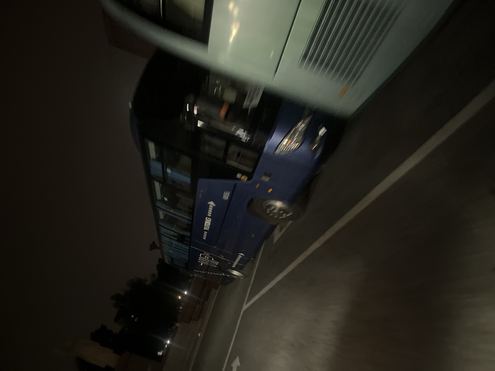 The image captures a night scene with two buses in motion, suggesting a travel or transportation context. The primary subject is a dark blue Yutong bus, partially obscured by a lighter colored bus to its right. The Yutong bus displays text and a logo on its side, possibly indicating its brand or purpose. The surrounding environment is dimly lit, with streetlights or other artificial light sources casting faint illumination on the asphalt road and parking lot markings. The motion blur in the image indicates that the buses are moving, and the shot was likely taken from a moving vehicle or by hand with a slow shutter speed. The overall atmosphere is urban and nocturnal.