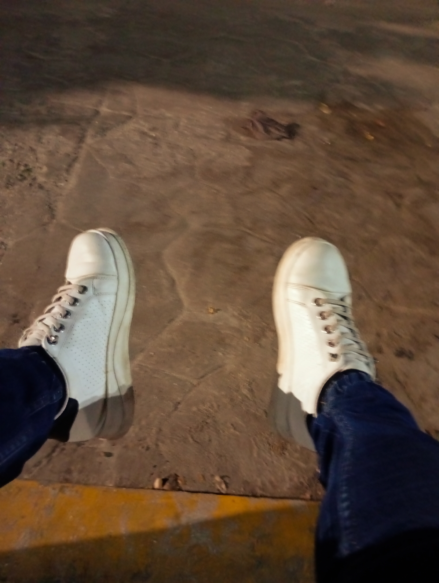 A person's legs are seen from a downward angle, with white sneakers and blue jeans resting on a weathered stone surface. The lighting is dim, suggesting an evening or nighttime setting. A yellow curb is visible in the lower left corner. The location is likely a public area in Juan Griego, Venezuela, given the information provided.