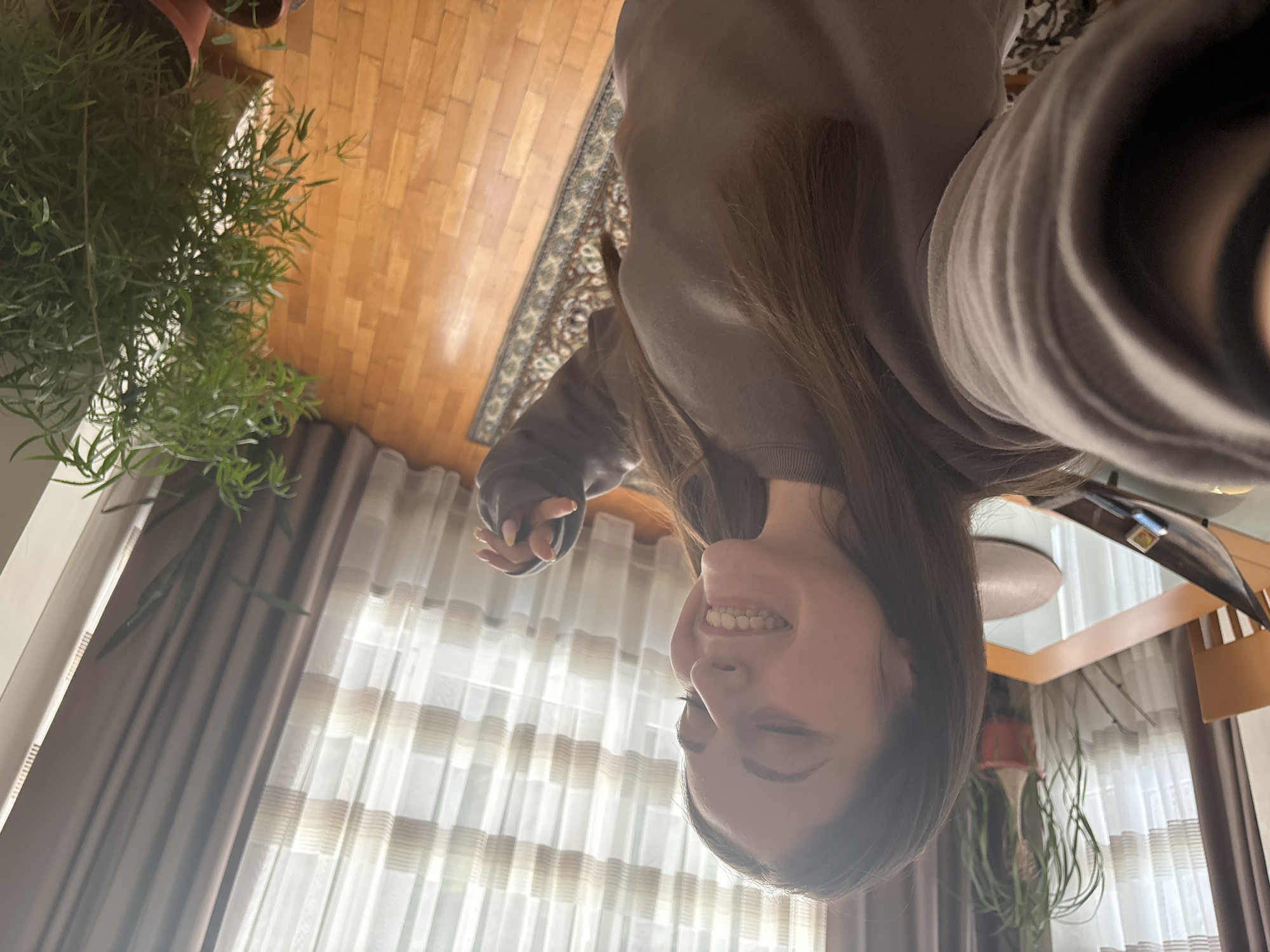 A brightly lit indoor selfie features a smiling young woman with long brown hair, wearing a dark gray or brown long-sleeved top. Her face, seen from a low angle, is cheerful, and she displays a broad smile, showing her teeth. Her right hand, partially curled, reveals light-colored fingernails.
The setting appears to be a living or dining room in Bulgarovo, Bulgaria, filled with natural light streaming through a window covered by sheer, horizontally striped curtains and darker brown drapes. In the background, a polished wooden floor is visible, partially covered by a patterned rug with intricate designs. Several green potted plants add to the decor; a large, leafy plant with slender fronds is prominent on the left side of the frame, and another smaller plant is visible near the bottom right. A wooden table with a reflective surface, possibly glass, is also present, reflecting parts of the room or a light fixture. The scene conveys a pleasant, homey atmosphere during daytime. No text is visible in the image.