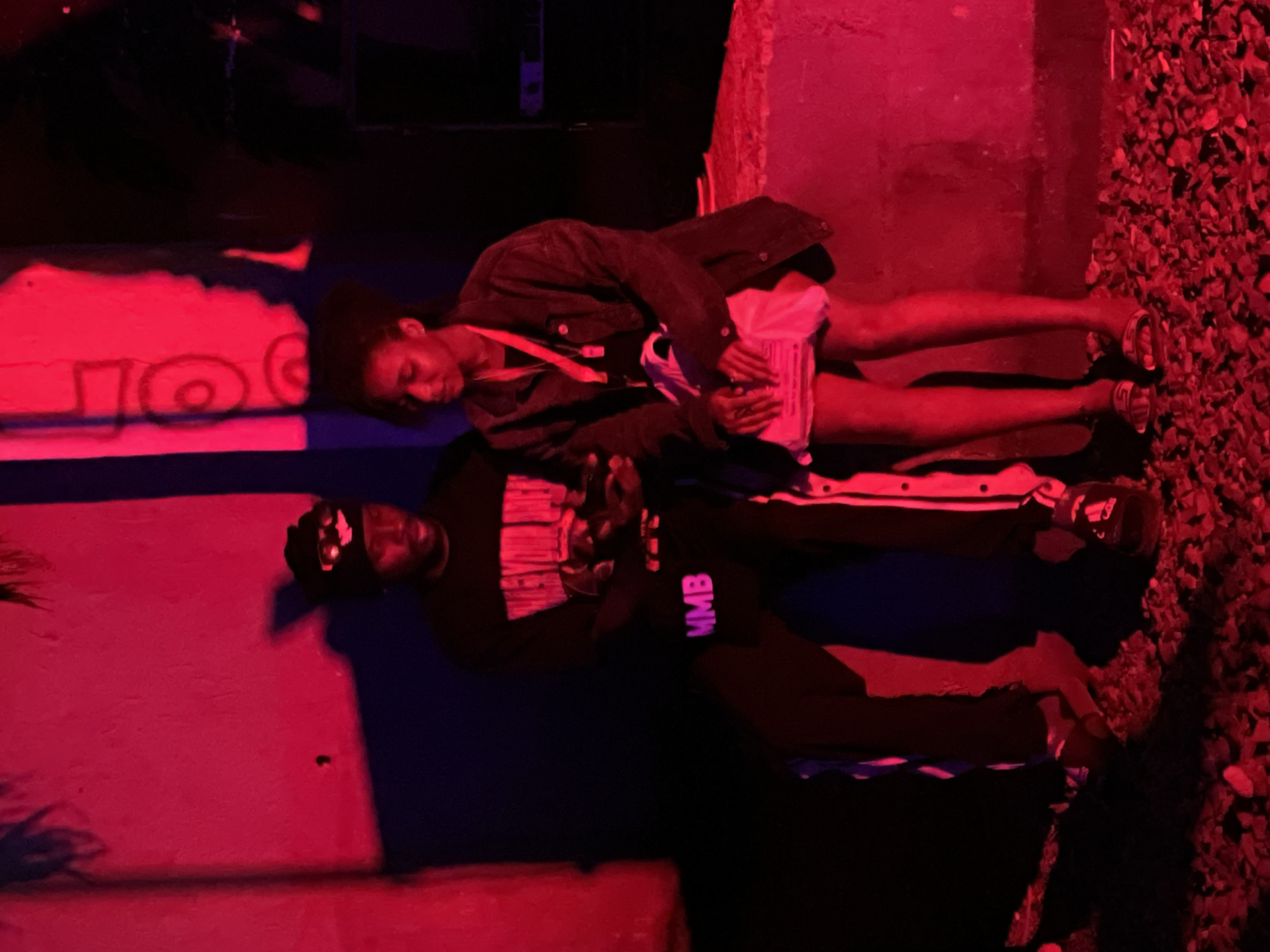 In Durumi, Nigeria, two individuals are captured sitting on a ledge under vibrant red lighting. The person on the left, a male, wears a black beanie with goggles and a black t-shirt with white lettering, and black pants with white stripes. The person on the right, a female, is dressed in a dark jacket over a black top, a pink lanyard with a white object attached, and a skirt, holding a white bag. The environment appears to be outdoors, with a textured ground beneath them and a wall behind. Shadows are cast dramatically by the red illumination, creating a moody atmosphere.