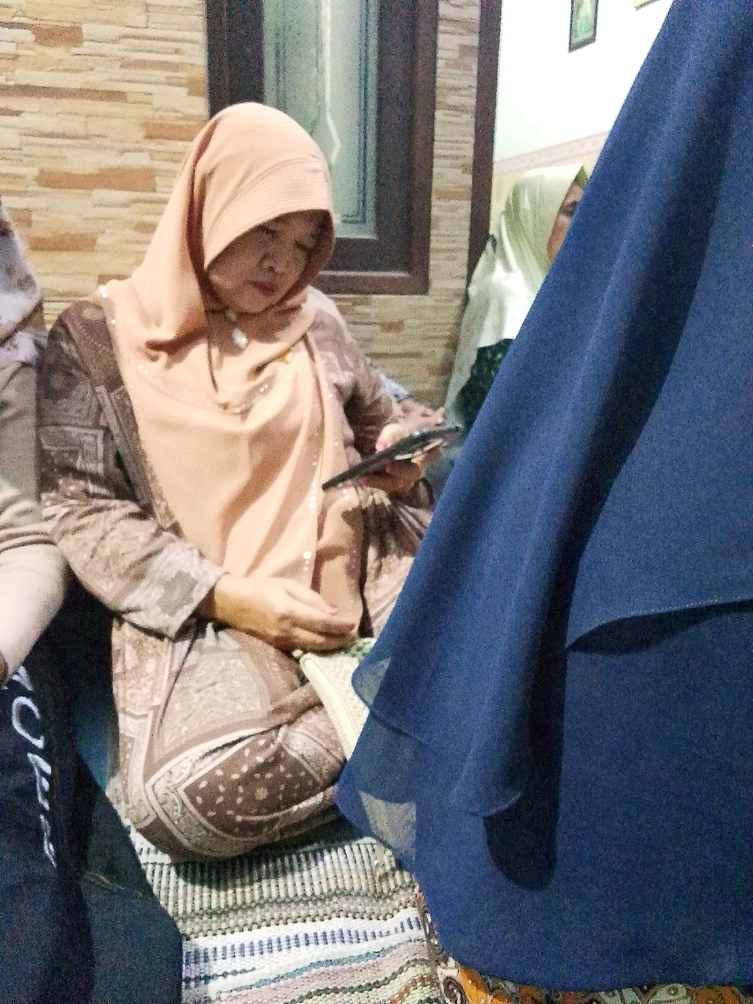 A woman in a peach-colored hijab and a patterned brown robe is seated on a woven mat. She is looking down and holding a black smartphone in her right hand. The background shows a textured wall and a window with a wooden frame. Another woman in a light green hijab is partially visible to her right. A large dark blue fabric is in the foreground, obscuring part of the scene. The setting appears to be an indoor residential area in Bulurejo, Indonesia.