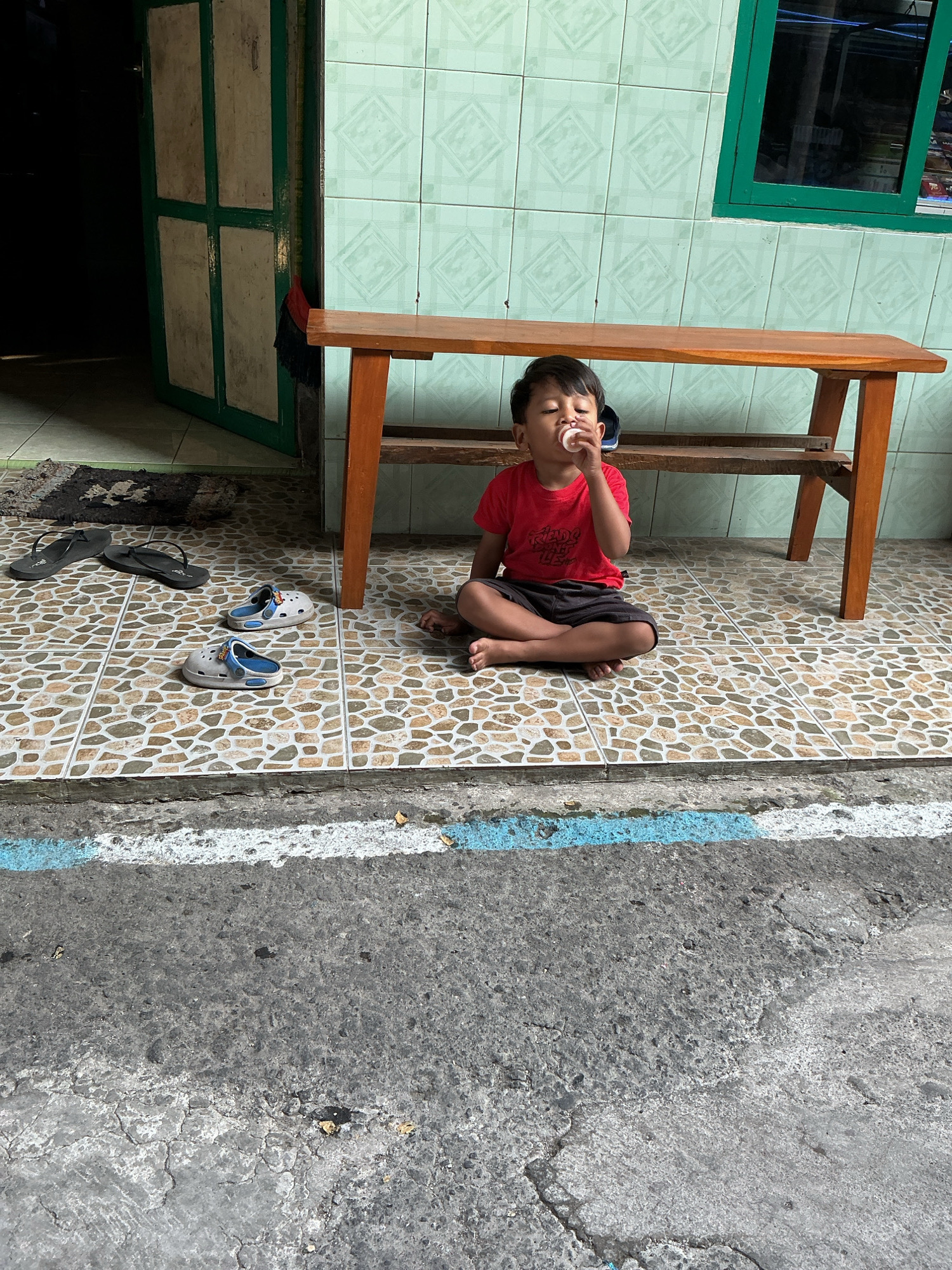In Magelang, Indonesia, a young child, possibly a boy, sits cross-legged on a patterned tiled floor in front of a wooden bench. The child wears a red t-shirt and dark shorts and is holding a small, white object to their mouth, appearing to suck on it. To the child's left are two pairs of footwear: a pair of black flip-flops and two pairs of white and blue sandals. Behind the child, a wooden bench with a slatted lower shelf is positioned against a wall tiled with light green diamond patterns. To the left of the bench, a green-framed door with white panels is partially open, revealing a dark interior. The foreground shows a cracked asphalt surface with a white and blue painted line. The scene appears to be an outdoor area of a residential building.