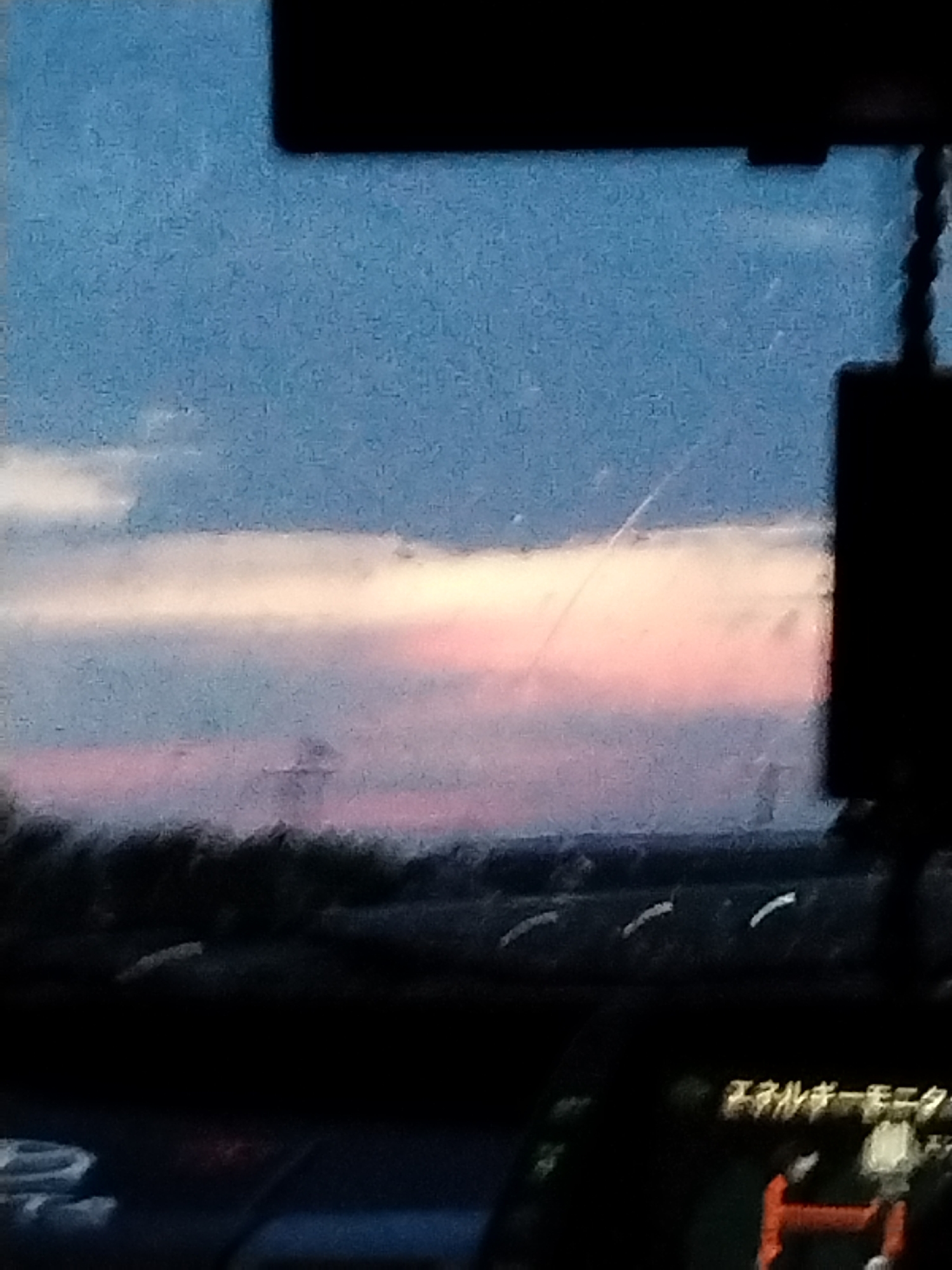 The image captures a blurry, low-light view of a sunset sky from within a vehicle. The sky is a gradient of blue, white, and pink, with some clouds visible. The horizon is obscured by a dark, out-of-focus landscape, likely trees or buildings. In the foreground, parts of the vehicle's interior are visible, including a dashboard with some blurry text in Japanese and a red object. The overall impression is one of a candid snapshot taken during twilight, with the specific location being Novonikol'sk, Russia, as indicated.
