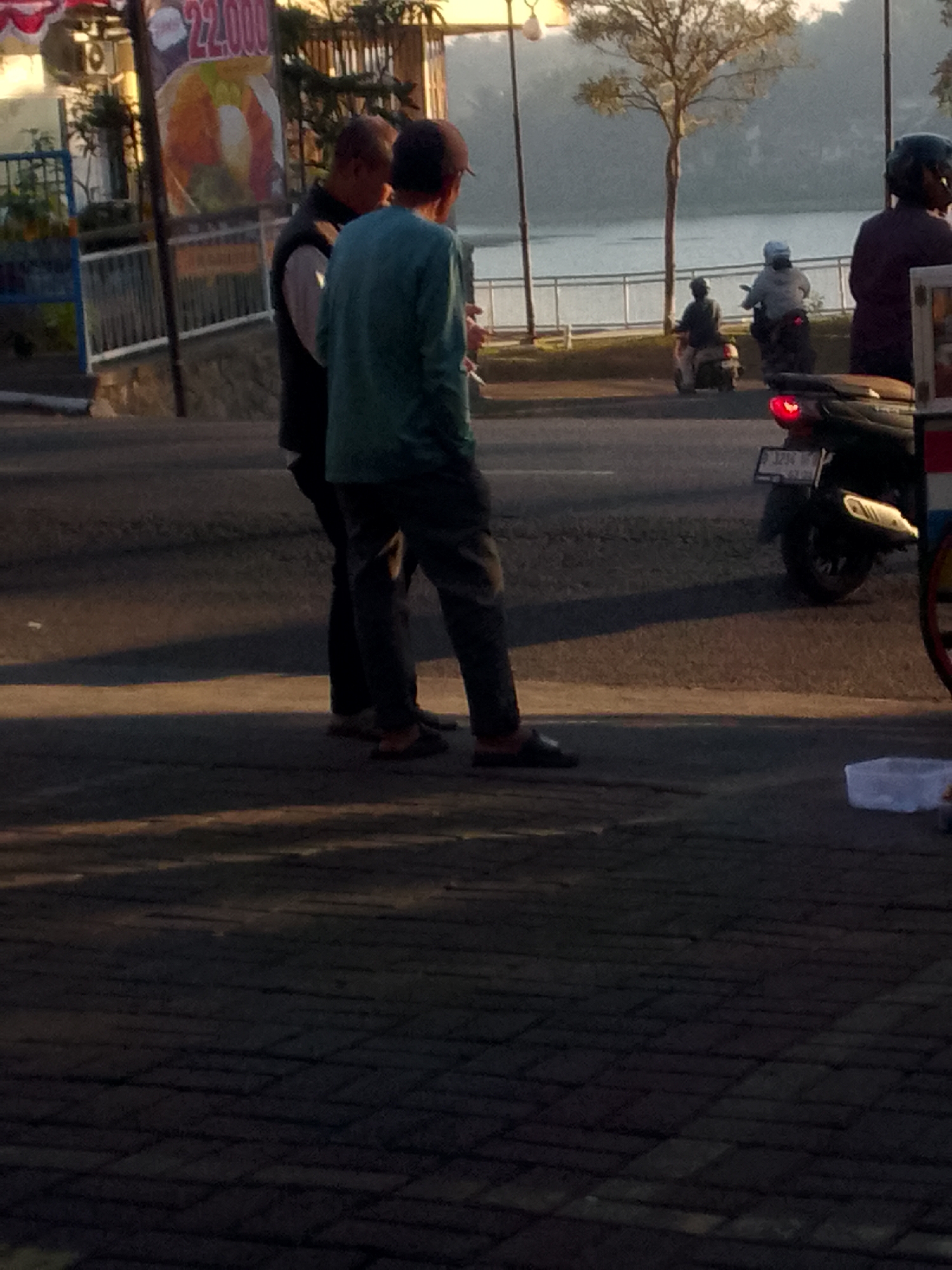 Two men stand on a paved surface in what appears to be an outdoor marketplace. The man on the left wears a dark vest over a white shirt and dark pants. The man on the right wears a teal long-sleeved shirt and dark pants. Both men are looking away from the camera. In the background, a body of water is visible, with a tree and a few distant figures on motorcycles. A sign with the number "22,000" is also visible in the background, along with a motorcycle with a red taillight. The scene suggests an early morning setting due to the lighting and the presence of mist over the water. The image does not contain specific landmarks to pinpoint the exact location in Ciburuy, Indonesia.