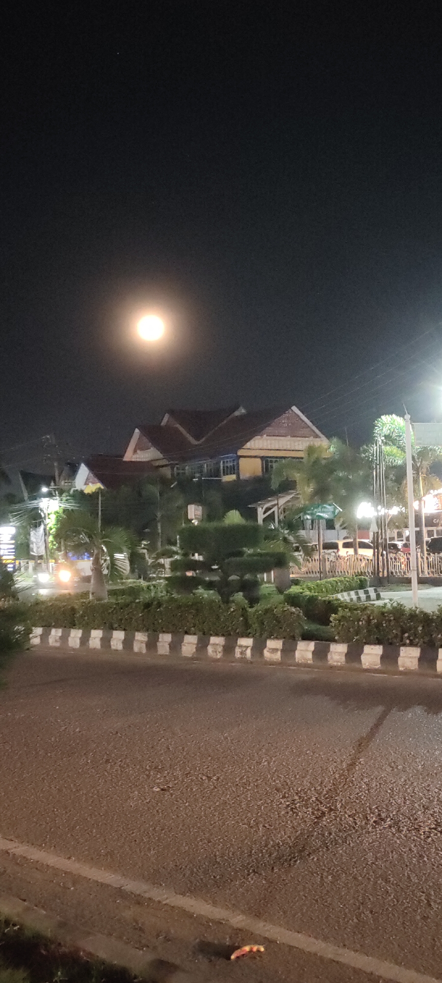 The image depicts a night scene in an unknown city. A full moon is visible in the sky above a building with a red-tiled roof and yellow walls. The building is surrounded by trees and manicured shrubbery, bordered by a black-and-white striped curb. A street runs in front of the curb, and streetlights illuminate the scene. A vehicle's headlights are visible in the distance.