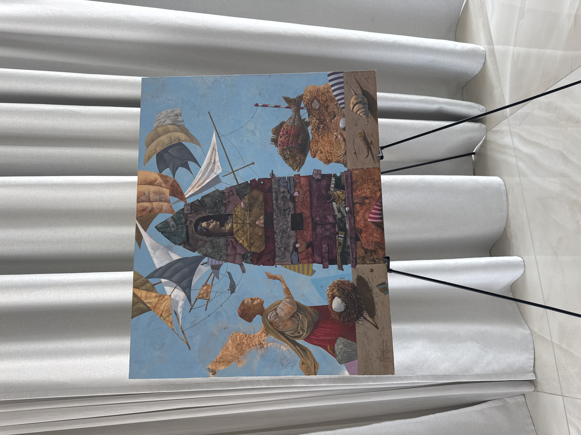 A surrealist painting is displayed on a black easel in front of a light grey curtain. The painting depicts a tall, multi-layered tower-like structure with a woman's face peering out from a window near the top. Sails of various colors and patterns fill the blue sky above the tower. To the left, a winged figure with golden wings and a red robe extends a hand towards a nest containing two white eggs. To the right of the tower, a large, intricately detailed fish rests on an orange rocky outcrop. The foreground of the painting shows a sandy ground with shells and a striped pattern. The bottom of the painting has a signature and a date, "Angel 2008". The scene is set indoors, likely in a gallery or exhibition space, with the floor appearing to be polished light marble.