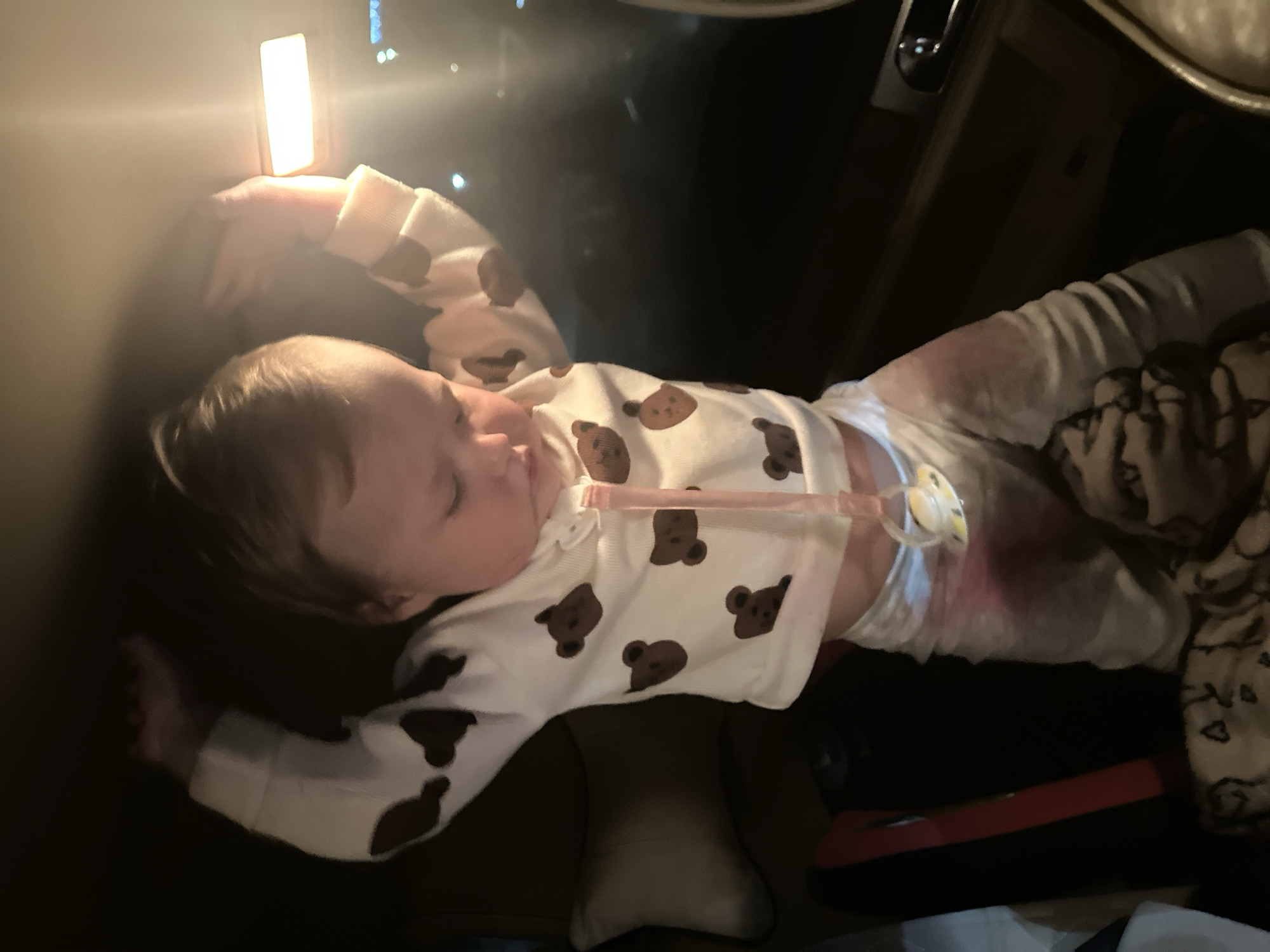 A baby is lying down, possibly sleeping, inside a vehicle. The child is wearing a white onesie with brown bear faces, a diaper, and has a pacifier attached by a strap. The vehicle's interior light is on. The location of Nizhny Novgorod, Russia, is not discernible from the provided image.