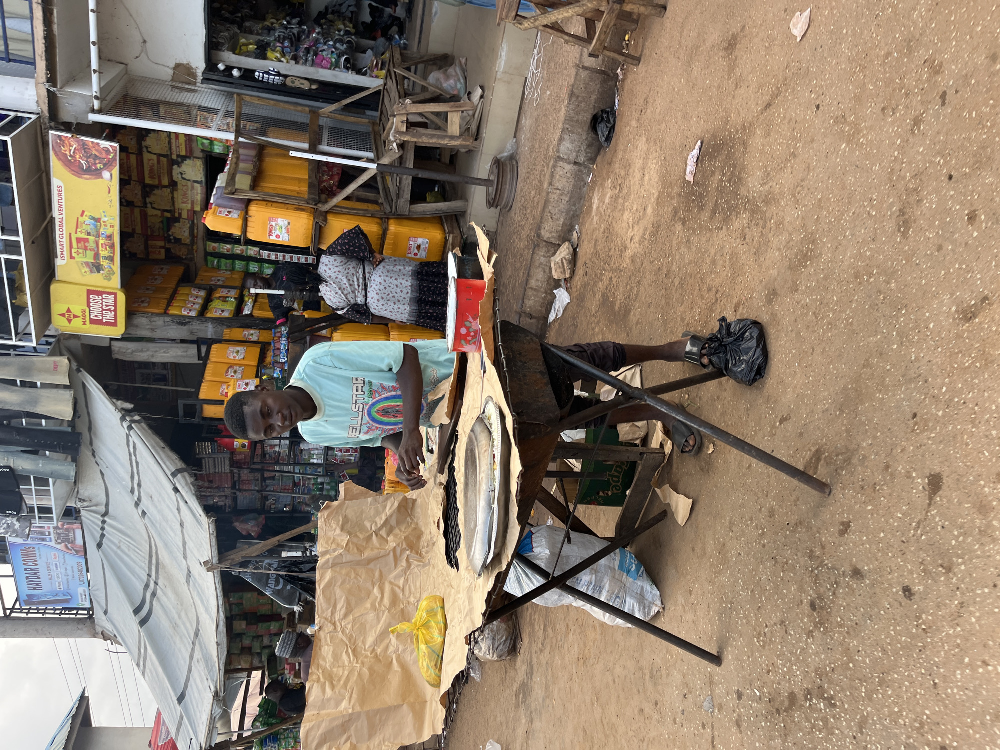A young man is seen in a marketplace in Bukuru, Nigeria, preparing fish for sale. He is standing behind a makeshift table made of brown paper and a metal frame, with a metal tray holding a whole fish. The man is wearing a light blue t-shirt with graphics and text, some of which reads "ELLSTAR". He appears focused on his task.
The scene is set in a bustling outdoor market. Shelves stocked with various goods, including what appear to be food items and cooking oil in large yellow containers, are visible in the background. Signs with text such as "HAYDAR COMMS", "MAGGI", and "KINGS" are displayed on the stalls. A woman in a patterned dress is also visible in the background. The ground is a mix of dirt and concrete, with some scattered debris. The overall impression is of a typical day in a Nigerian marketplace, with vendors selling their wares. The lighting suggests it could be daytime, possibly mid-morning or afternoon.