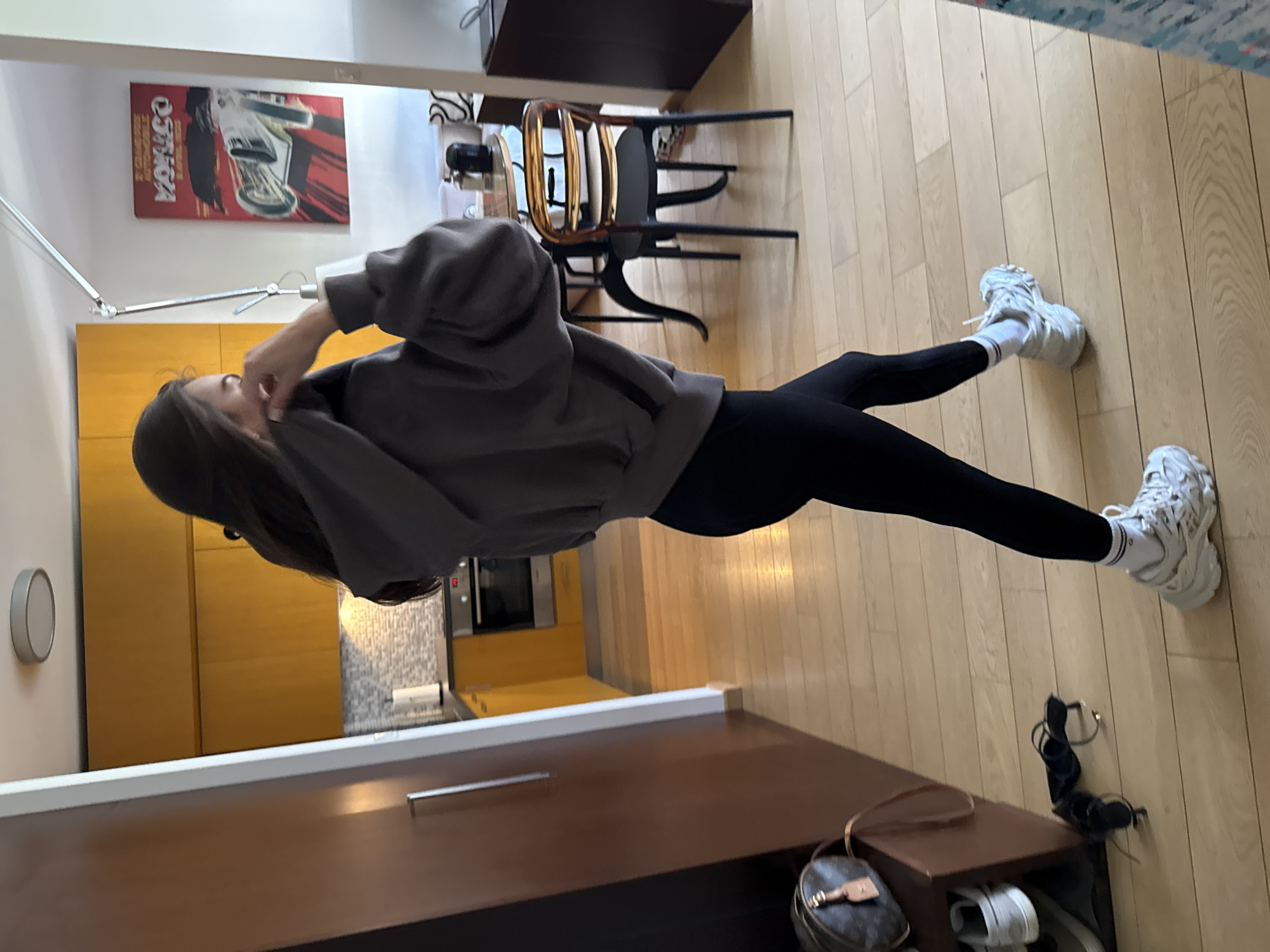 A woman wearing a gray hooded sweatshirt and black leggings is standing in a room with wooden floors. She is wearing white socks and sneakers. There is a table with a chair behind her, a cabinet in front of her, and a poster on the wall. The city of Unknown, Unknown, cannot be determined from the image.