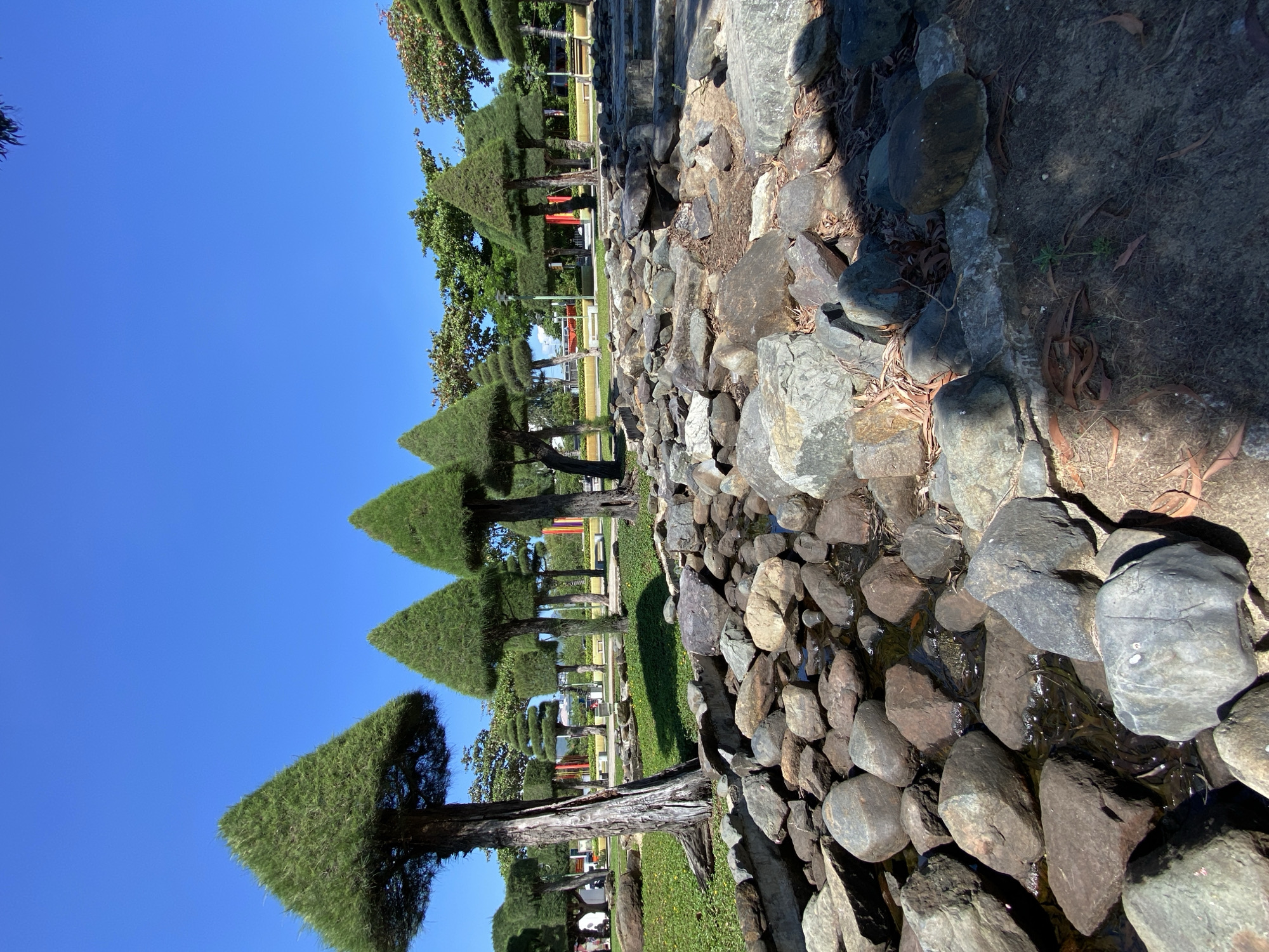 This image depicts a landscaped park with meticulously trimmed trees, some shaped into perfect cones, lining a path. The scene is characterized by a clear, bright blue sky, suggesting a sunny day. In the foreground, a rocky stream bed is visible, filled with various sized stones and a shallow flow of water. Sunlight casts shadows on the rocks, indicating it is likely midday.
In the background, more trees and foliage are apparent, along with what appear to be park benches or seating areas. Several red structures are also visible in the distance, adding pops of color to the green and blue landscape. The overall impression is one of a well-maintained, peaceful outdoor space, possibly a garden or a public park. The groomed trees and the water feature suggest a deliberate design aimed at aesthetic appeal. The location context of Nha Trang, Vietnam, implies a tropical or subtropical climate, consistent with the lush greenery and clear sky. There is no visible text in the image.