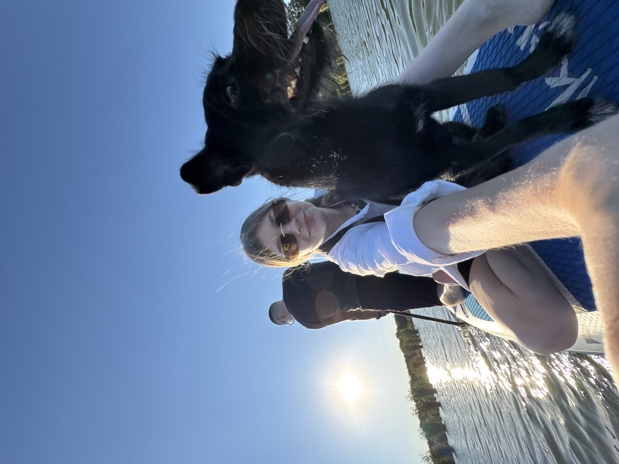 This is a selfie taken from a paddleboard on a body of water under a clear blue sky. The sun is setting or rising, casting a bright glare on the water and behind a person standing on the paddleboard. A woman is in the foreground, holding the camera and smiling. Next to her is a black dog, looking towards the camera with its mouth open. Another person is standing on the paddleboard in the background, holding a paddle. The paddleboard is blue and white. Trees are visible in the distance along the shoreline. The location is identified as Novotitarovskaya, Russia.