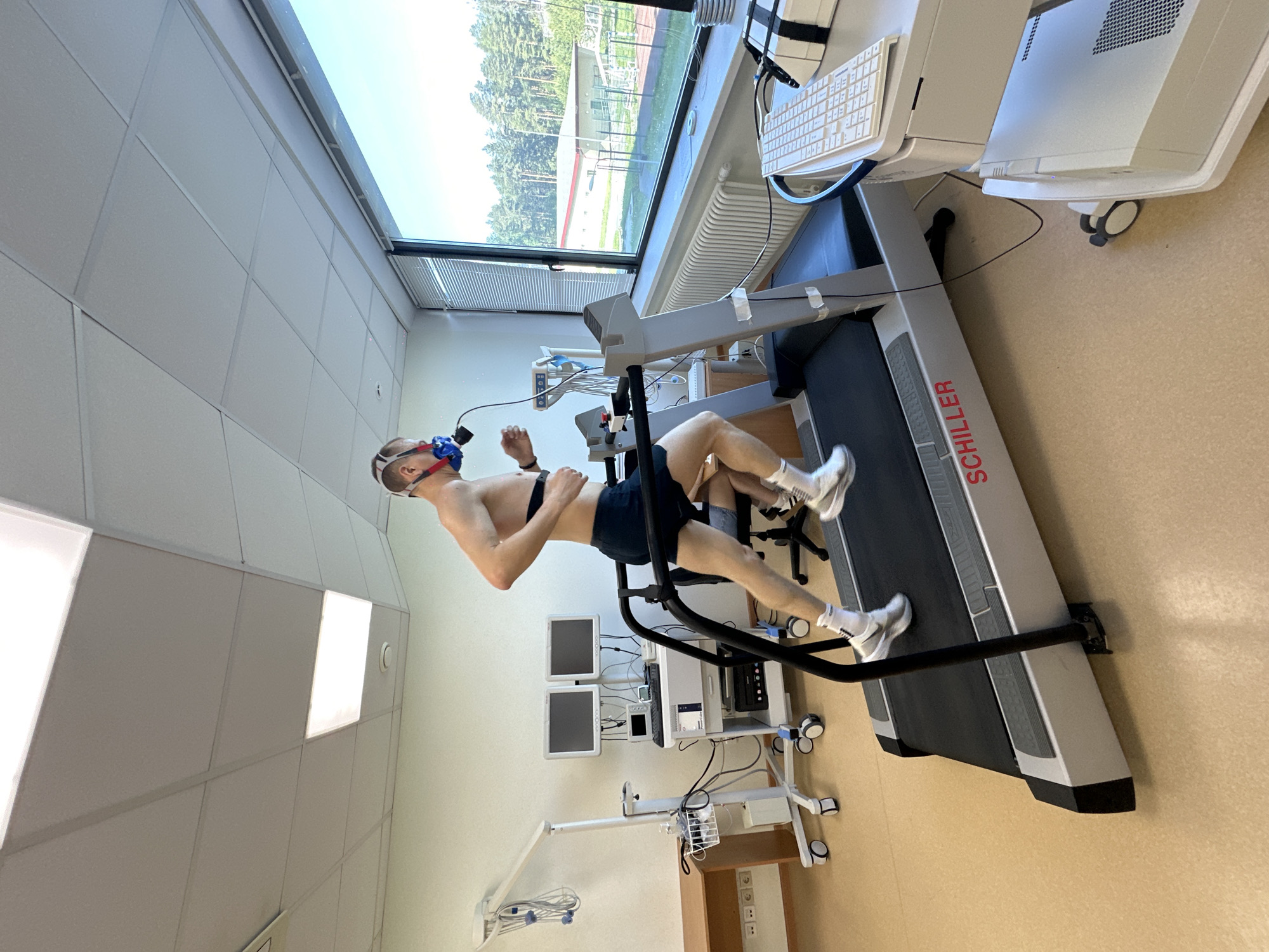 A man with a mask on his face is running on a Schiller treadmill in a medical examination room. The room has white walls and a large window. In the background, trees and a building can be seen, suggesting an exterior view. A medical device cart with monitors is beside the treadmill. The location might be Прикамский, Russia, given the user query, but this can't be confirmed from the image alone.