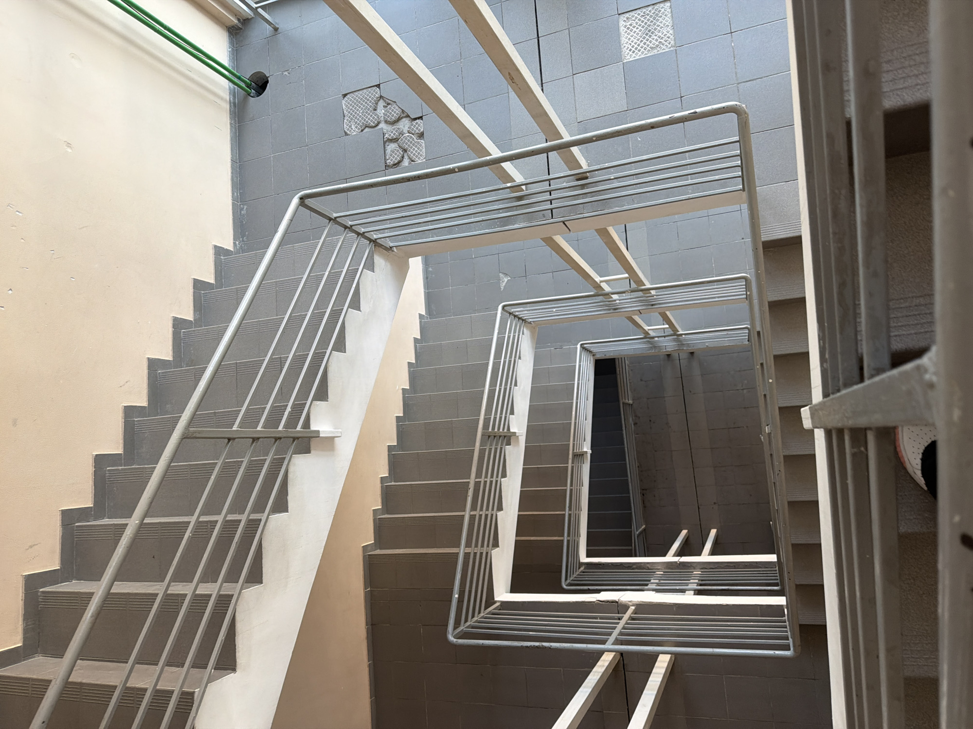 An interior shot of a stairwell looks down from a high angle. The stairwell is constructed with concrete steps and grey tiled floors. A silver metal railing runs along the sides of the stairs and the perimeter of the stairwell. The walls are painted a neutral beige color. The lighting suggests it is daytime, possibly indoors with artificial lighting. There are no visible people. The image was taken in Kyiv, Ukraine.