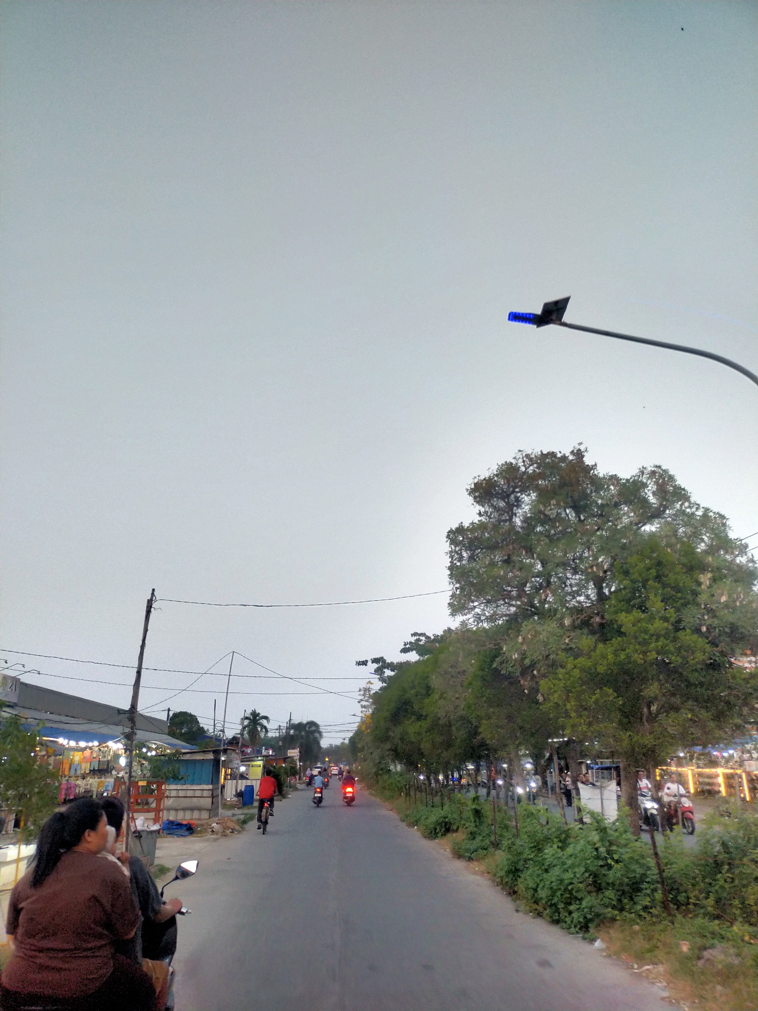 The image depicts a street scene in Babakan Asem, Indonesia. The perspective appears to be from the back of a motorcycle, looking forward. On the left side of the road, there are several stalls with blue awnings, displaying various goods, possibly clothing. A person in a brown shirt is seated on the back of the motorcycle in the foreground. To the right of the road, there is a line of trees with lush green foliage, some of which are bearing large, elongated pods. A street lamp with a blue light fixture is visible in the upper right portion of the frame. The road itself is paved and appears to be a two-lane street with moderate traffic. Several other motorcycles and a bicycle are on the road, moving in the same direction as the viewer. The sky is overcast and a pale grey. The overall atmosphere suggests it is late afternoon or early evening.