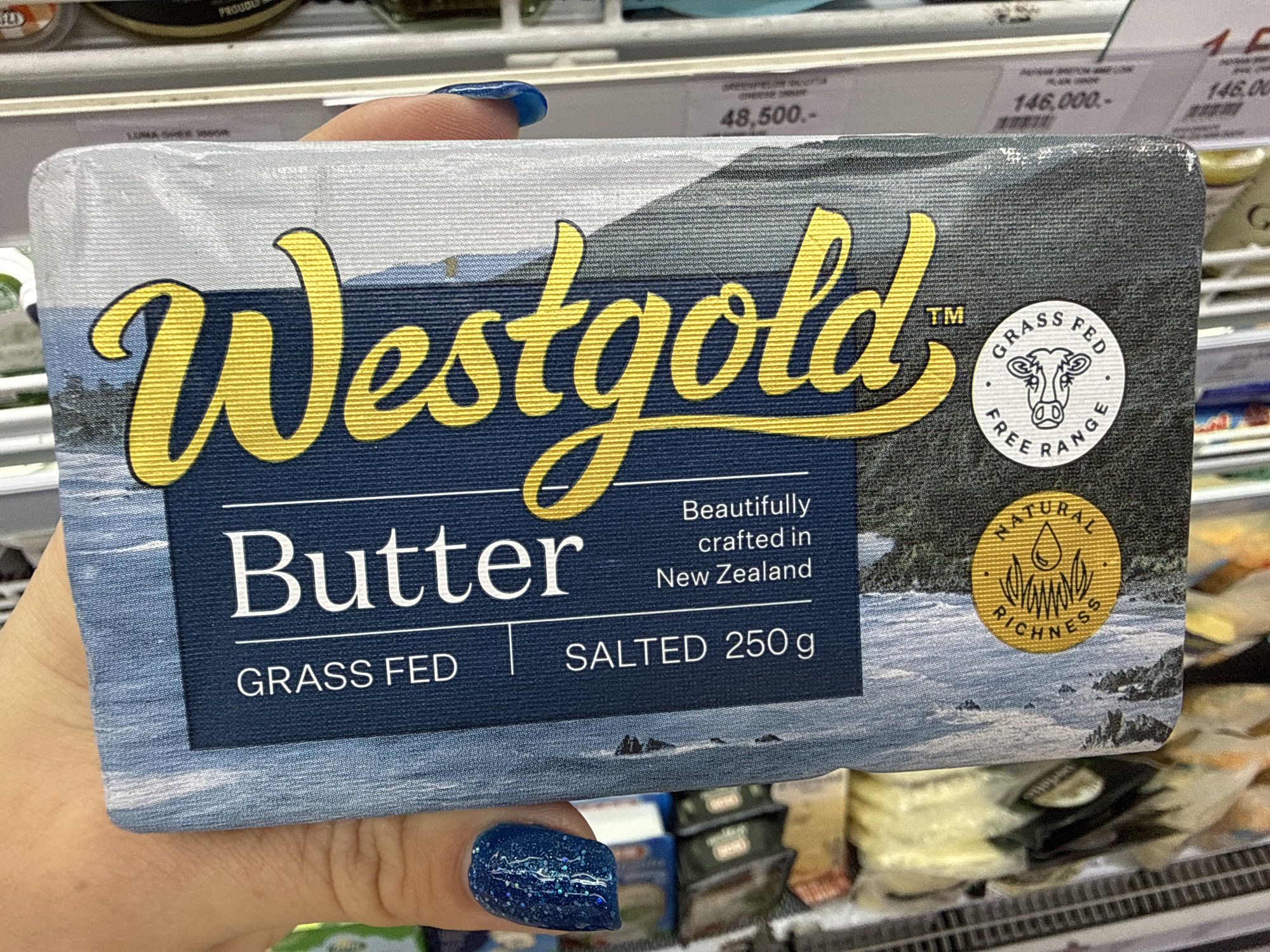 A person's hand with blue glitter nail polish is holding a package of "Westgold" butter. The butter package is a light blue color with a landscape image of mountains and trees. The text on the package is in yellow and white. It reads "Westgold Butter Grass Fed Salted 250 g Beautifully crafted in New Zealand." There are two circular seals on the package. One is yellow and says "Natural Riches." The other is white and says "Free Range Grass Fed" with a drawing of a cow's head. The background shows grocery store shelves with various products, including what appear to be other butter or dairy products. There are price tags visible on the shelves with Indonesian currency ("48.500.-" and "146.000.-"). The image is taken in a supermarket in Denpasar, Indonesia.