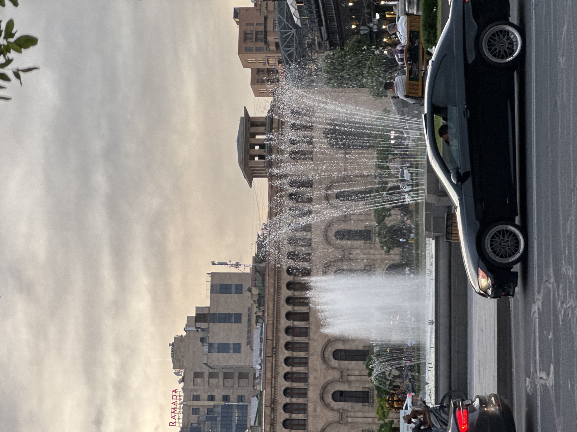 The image captures a scene in Yerevan, Armenia, featuring a fountain and surrounding buildings. The camera seems tilted 90 degrees. A large fountain dominates the center, shooting water jets into the air. Behind the fountain is a multi-story building with arched windows. To the left, a Ramada hotel sign is visible on another tall building. A black car is parked on the right side of the road, partially obstructing the view of the fountain. Other cars and pedestrians are seen in the background, near the base of the fountain. The sky is overcast.